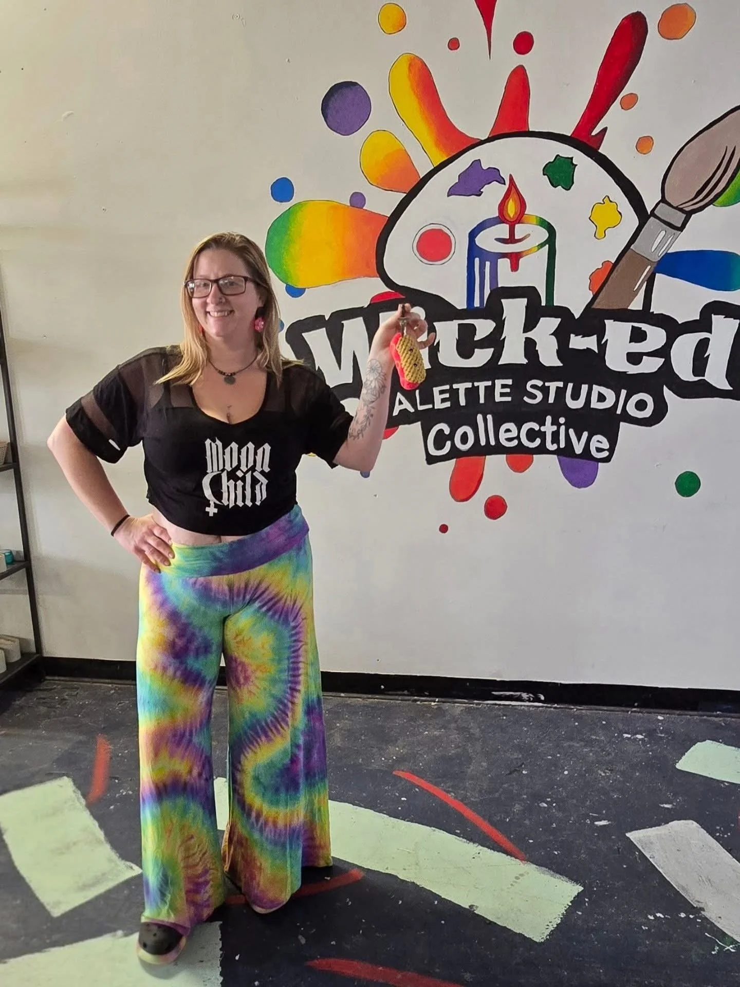 A woman with glasses and long hair smiling, wearing a black t-shirt with white text, tie-dye rainbow patterned wide-leg pants, holding a keychain, standing in front of a colorful art mural featuring a candle, paintbrush, and palette, with vibrant spl