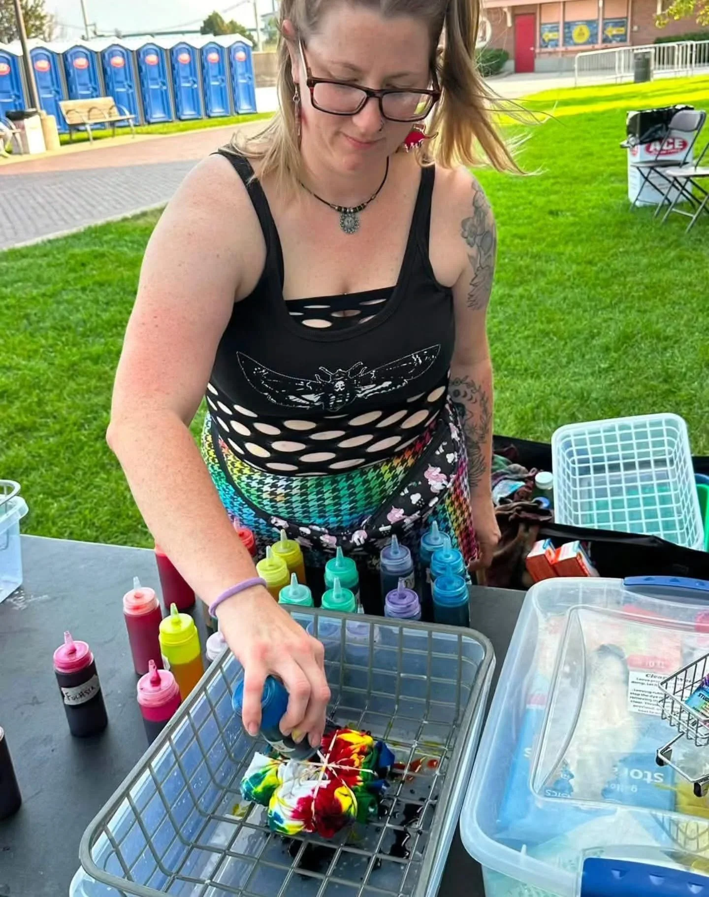 A woman wearing glasses and a black tank top with cut-outs, colorful patterned shorts, is sitting at a table outside, surrounded by bottles of colorful colored dye, and appears to be dyeing a tie-dye shirt or fabric. There are various containers and 