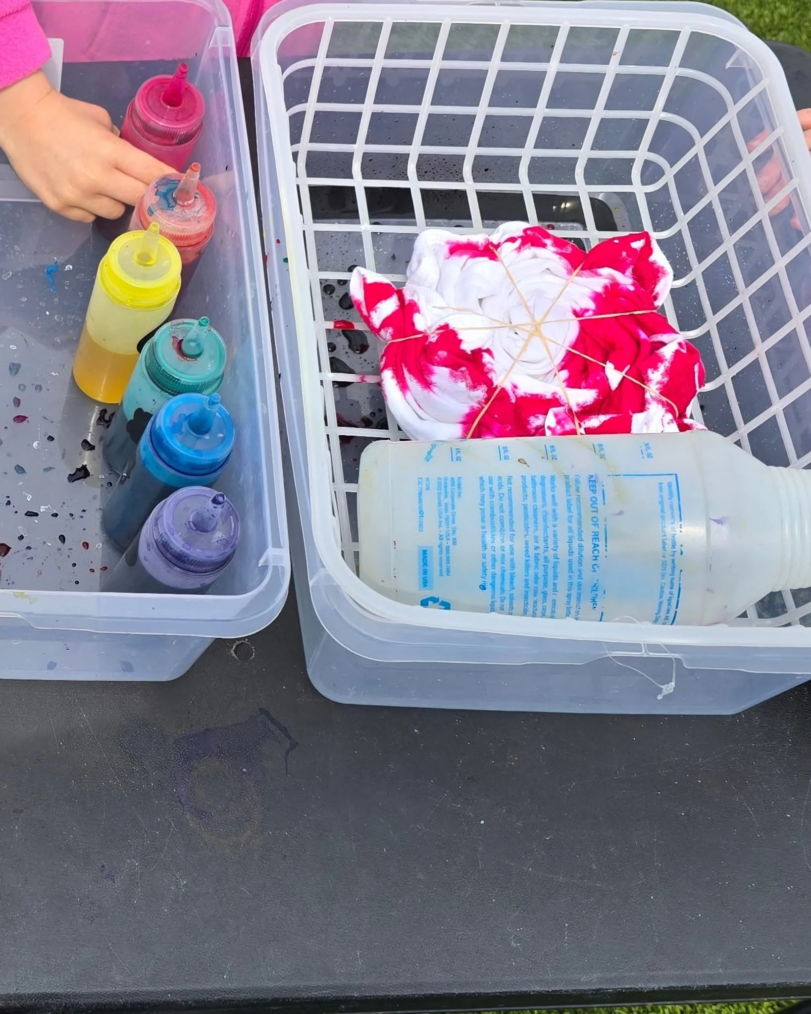 Plastic containers with paint bottles, a white plastic basket with a tie-dye cloth, and a plastic bottle are placed on a black table outdoors.