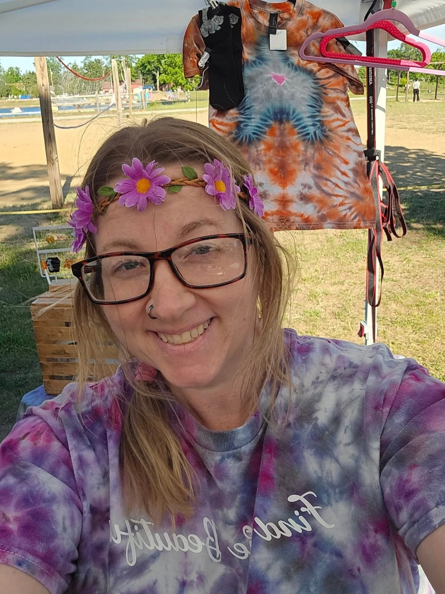 A young woman smiling outdoors, wearing glasses and a purple tie-dye shirt with the words 'Find the Beauty' written on it. She has a flower crown on her head and is taking a selfie.