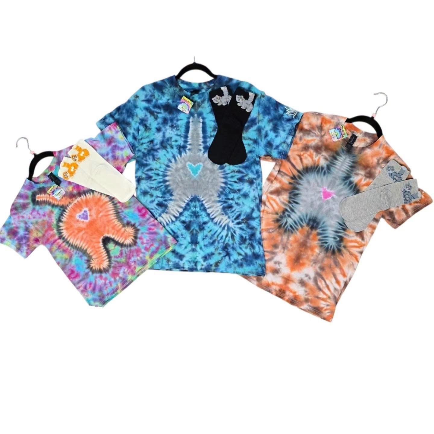 Three tie-dye T-shirts with cat graphic designs, paired with socks, displayed on black hangers against a white background.