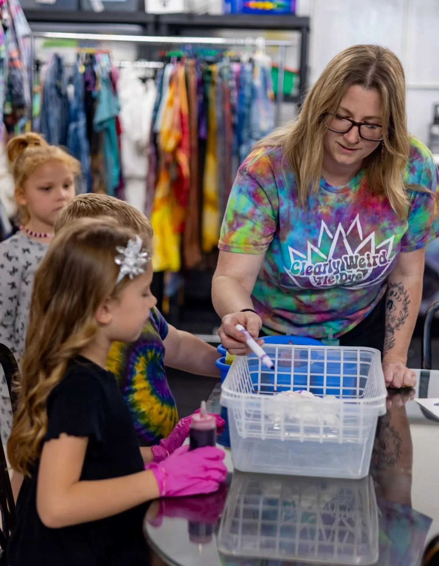A woman and three young girls with long hair, some with colorful tie-dye shirts and a snowflake hair clip, are gathered around a table. The woman is wearing a multicolored tie-dye shirt with the text "Clearly Weird" on it and glasses. They appear to 