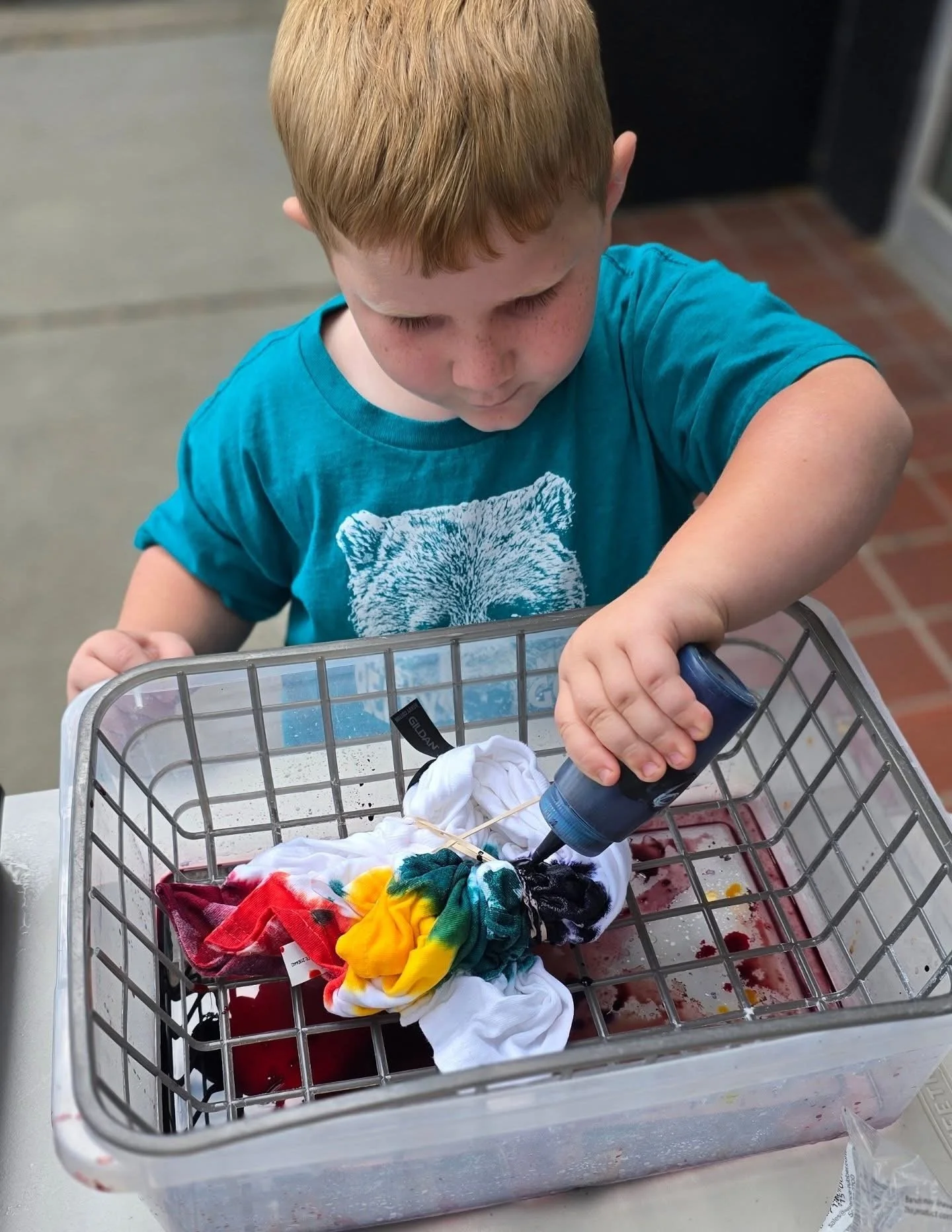 A young boy with red hair dyeing a white sock in a small shopping cart using a handheld dye spray bottle. There are colorful tie-dye cloths inside the cart.