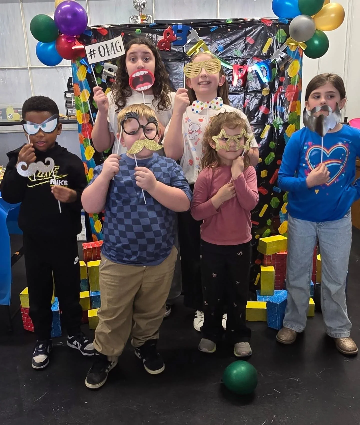 Group of children at a birthday party posing with fun photo props, including glasses, mustaches, lips, and a face mask, in front of a colorful decorated backdrop with balloons and gift boxes.
