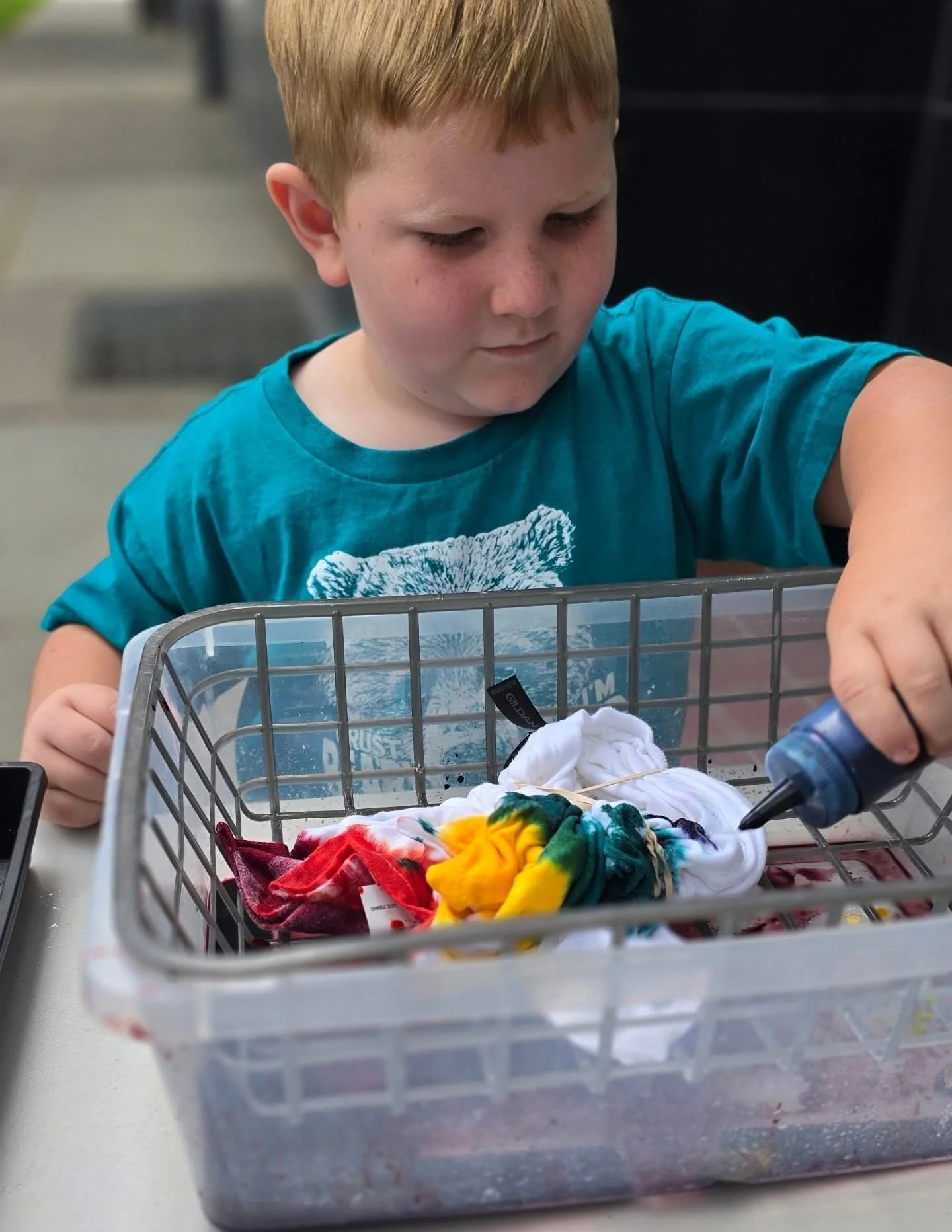 Young boy with red hair and a blue t-shirt, focused on tie-dyeing colorful fabric in a plastic basket at a table.