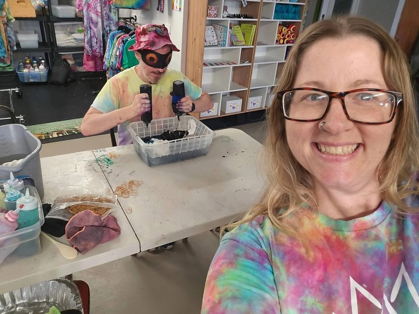 A woman with glasses and a nose piercing smiling for a selfie in the foreground, wearing a colorful tie-dye shirt. In the background, a man with a colorful hat, tie-dye shirt, and a mask, working with black dye on fabric at a table in a craft store.