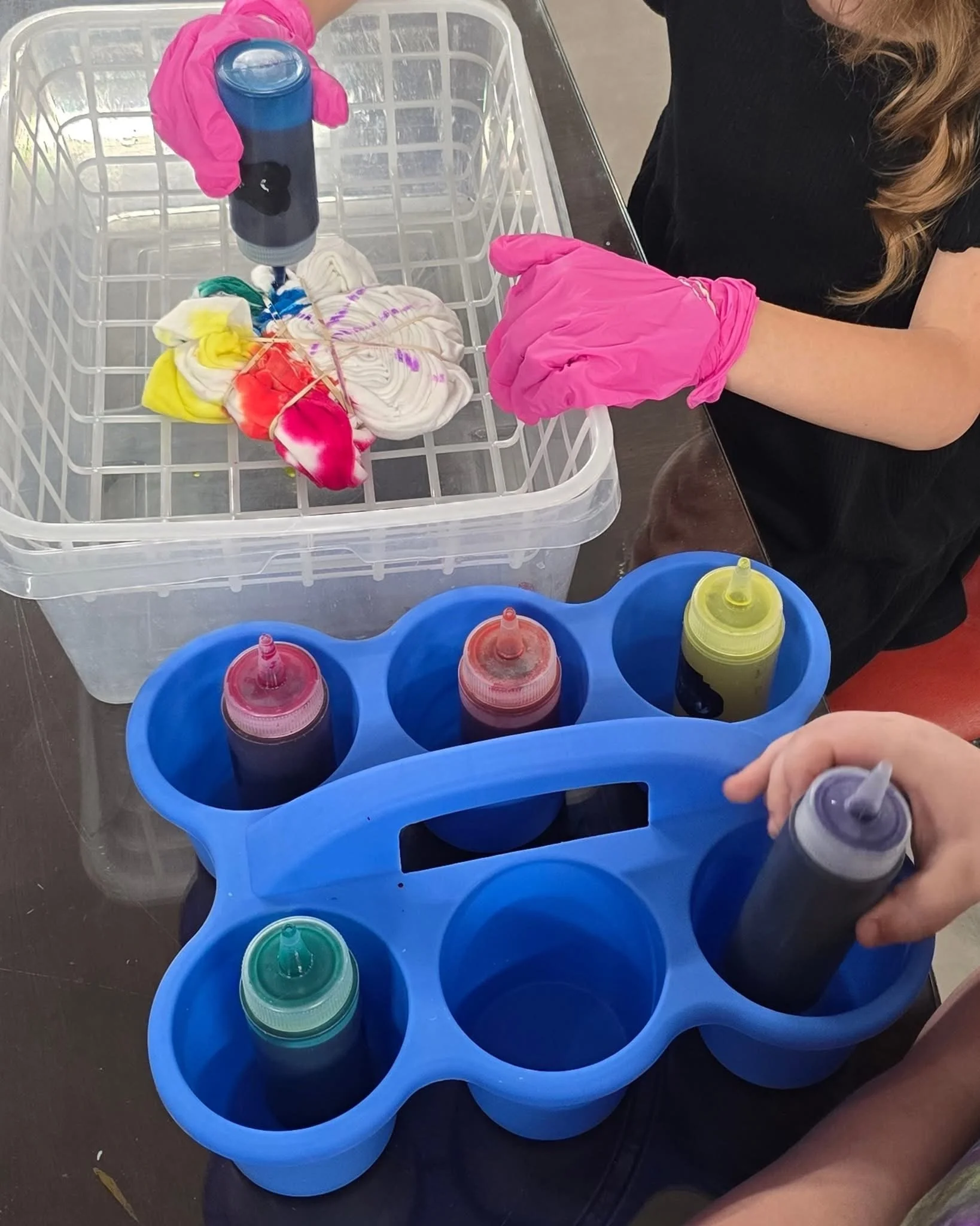 Person wearing pink gloves pouring colorful dye into small containers on a blue plastic tray, with a white container and colorful fabric scraps nearby.