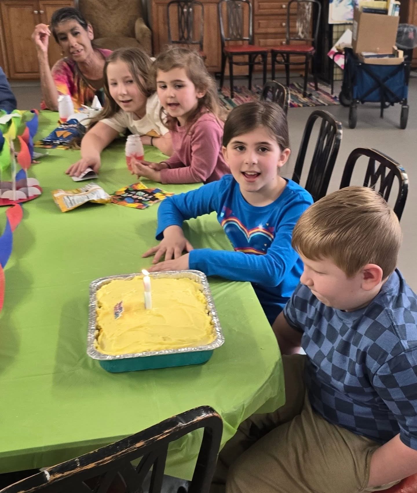 Children celebrating a birthday with a cake, a candle, and colorful decorations at a table covered with a green tablecloth.