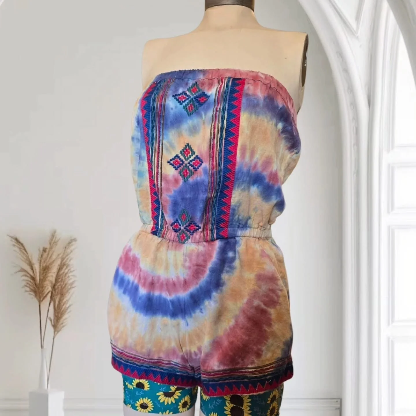 Back view of a mannequin wearing a strapless tie-dye dress with colorful embroidery patterns and a pair of blue shorts with sunflower print, standing indoors near a white wall and window, with dried pampas grass in a vase