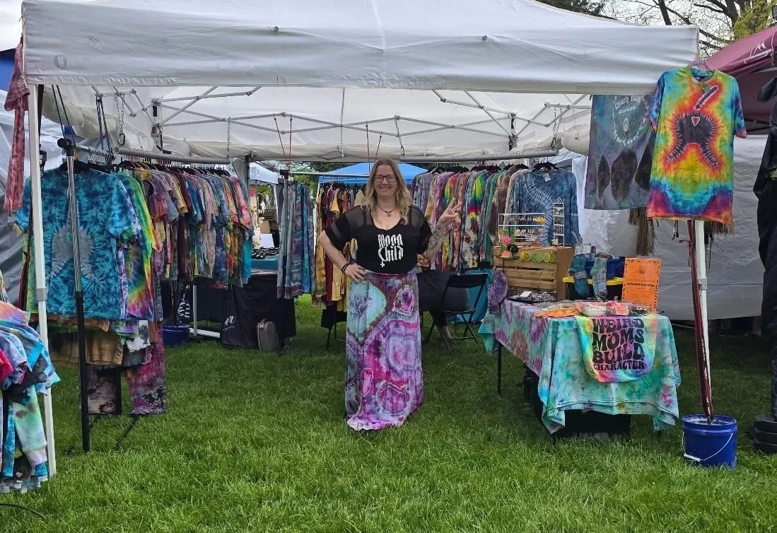 A woman standing inside a tie-dye clothing booth at an outdoor market, surrounded by racks of colorful tie-dye shirts and additional tie-dye merchandise on tables and hangers.