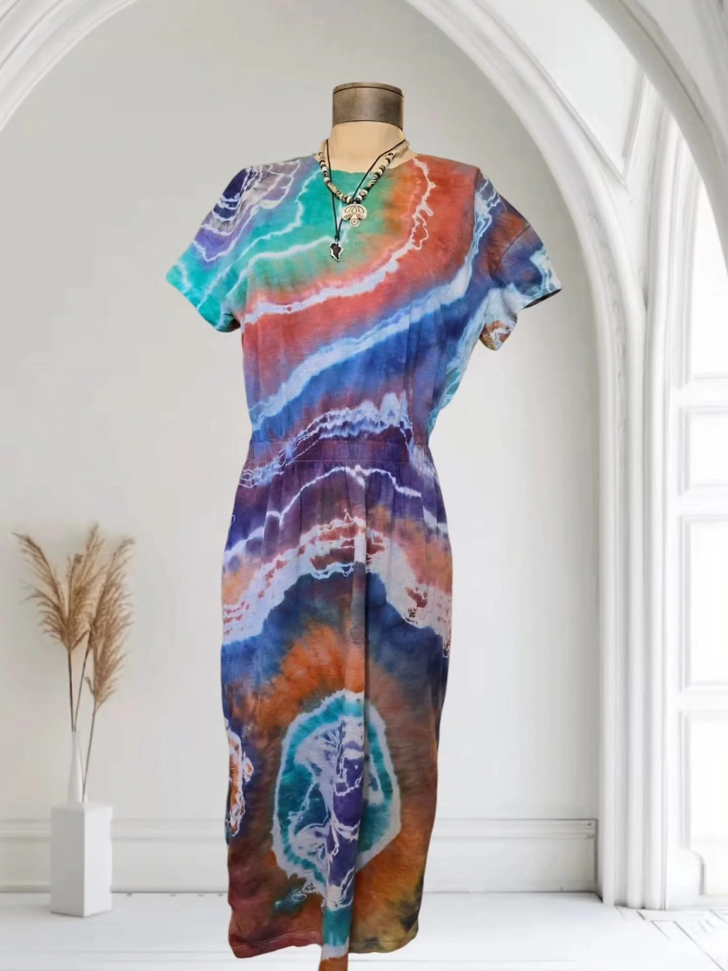 A mannequin wearing a colorful, tie-dye styled top and skirt, accessorized with layered necklaces, against a white wall inside a well-lit room.