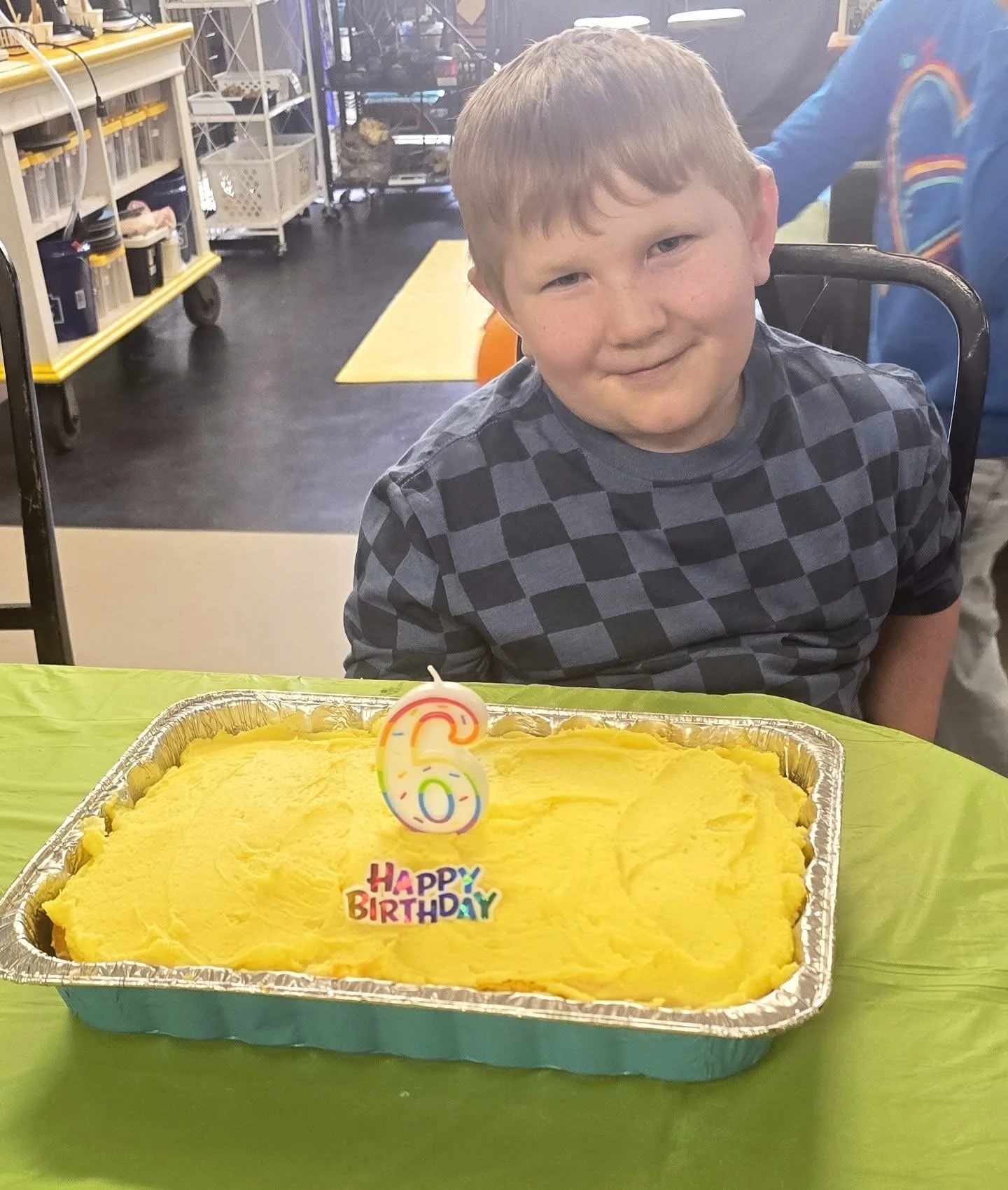 A young boy with short hair wearing a dark checkered shirt sitting at a table during his birthday celebration. In front of him is a yellow birthday cake with a colorful 'Happy Birthday' decoration and a number 6 candle. The boy has a slight smile and