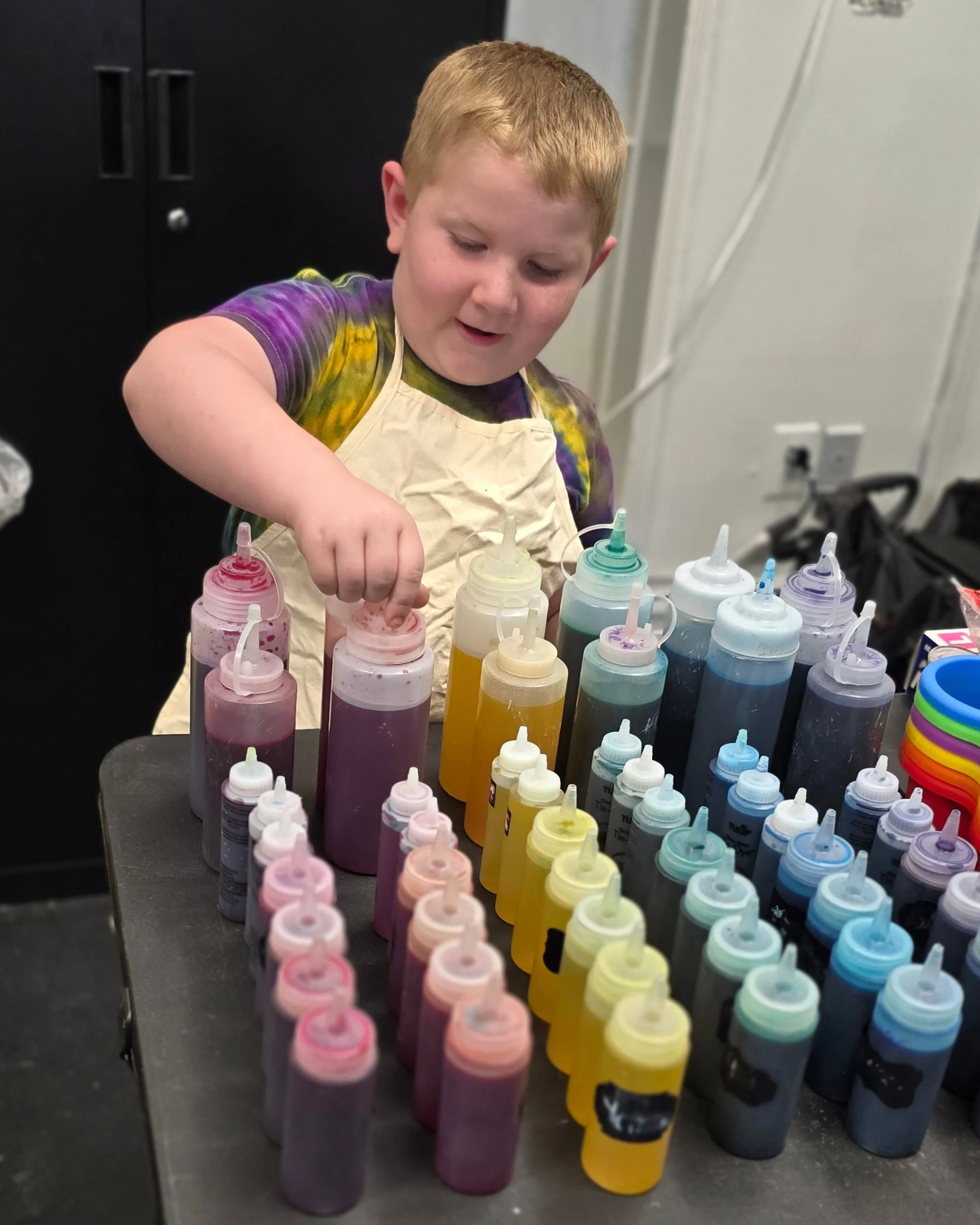 A young boy with red hair wearing a tie-dye shirt and apron, standing at a table with various bottles of colored paints, in a room with black cabinets and a white wall.