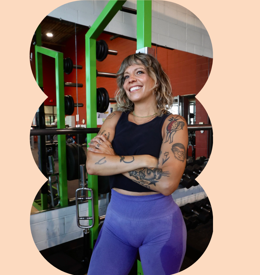 A woman with tattoos, wearing a black tank top and purple leggings, smiling with her arms crossed in a gym, standing in front of a weightlifting rack with green bars and black weights.