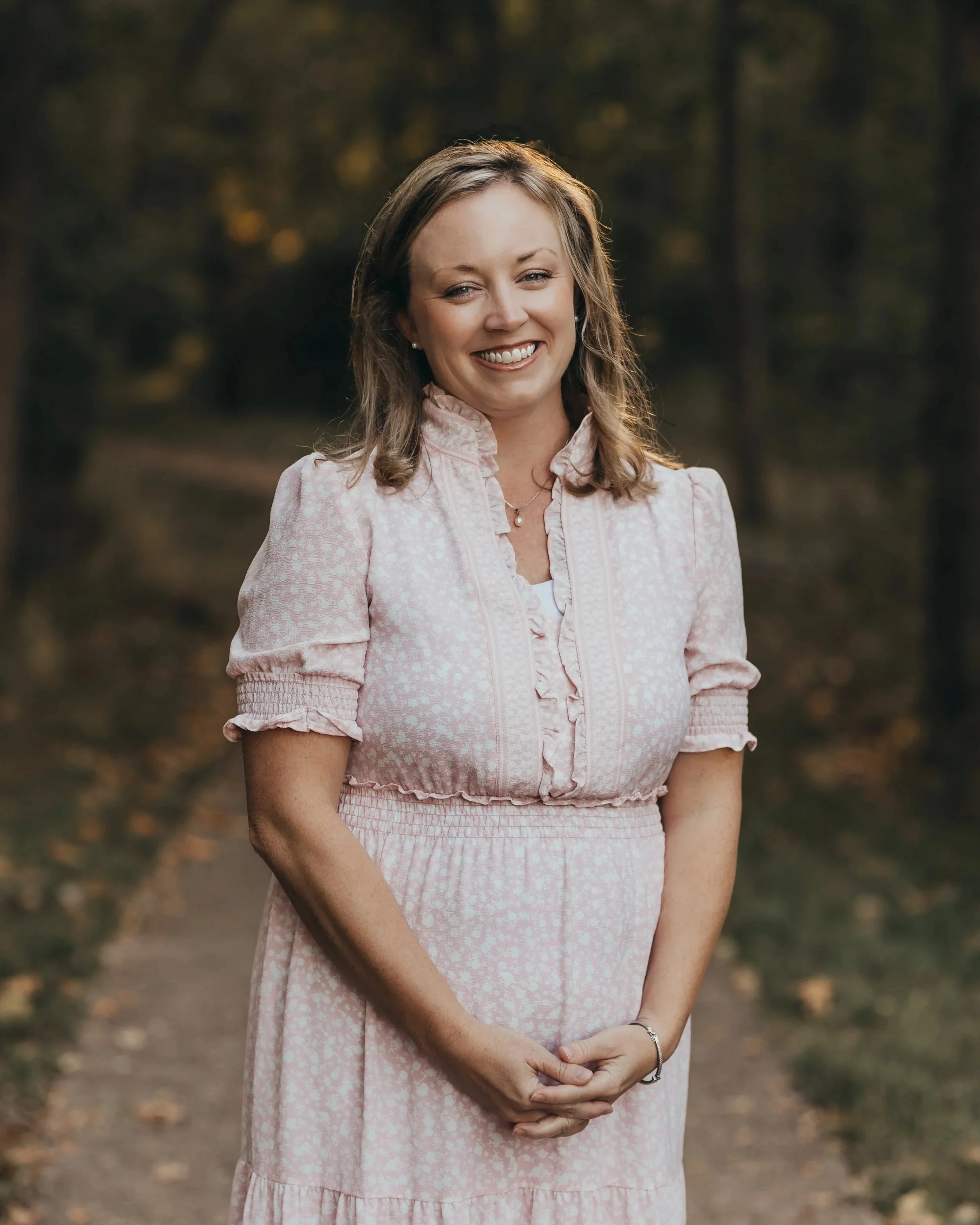 A woman with shoulder-length light brown hair, smiling, standing outdoors in a park-like setting during autumn. She is wearing a pink patterned dress with a ruffled collar and sleeves, and a silver bracelet.