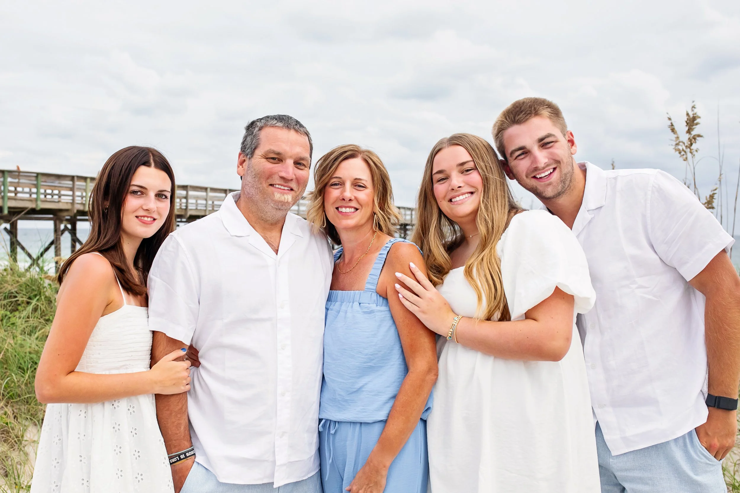 A group of five people smiling and posing outdoors near a wooden pier, with a cloudy sky in the background.