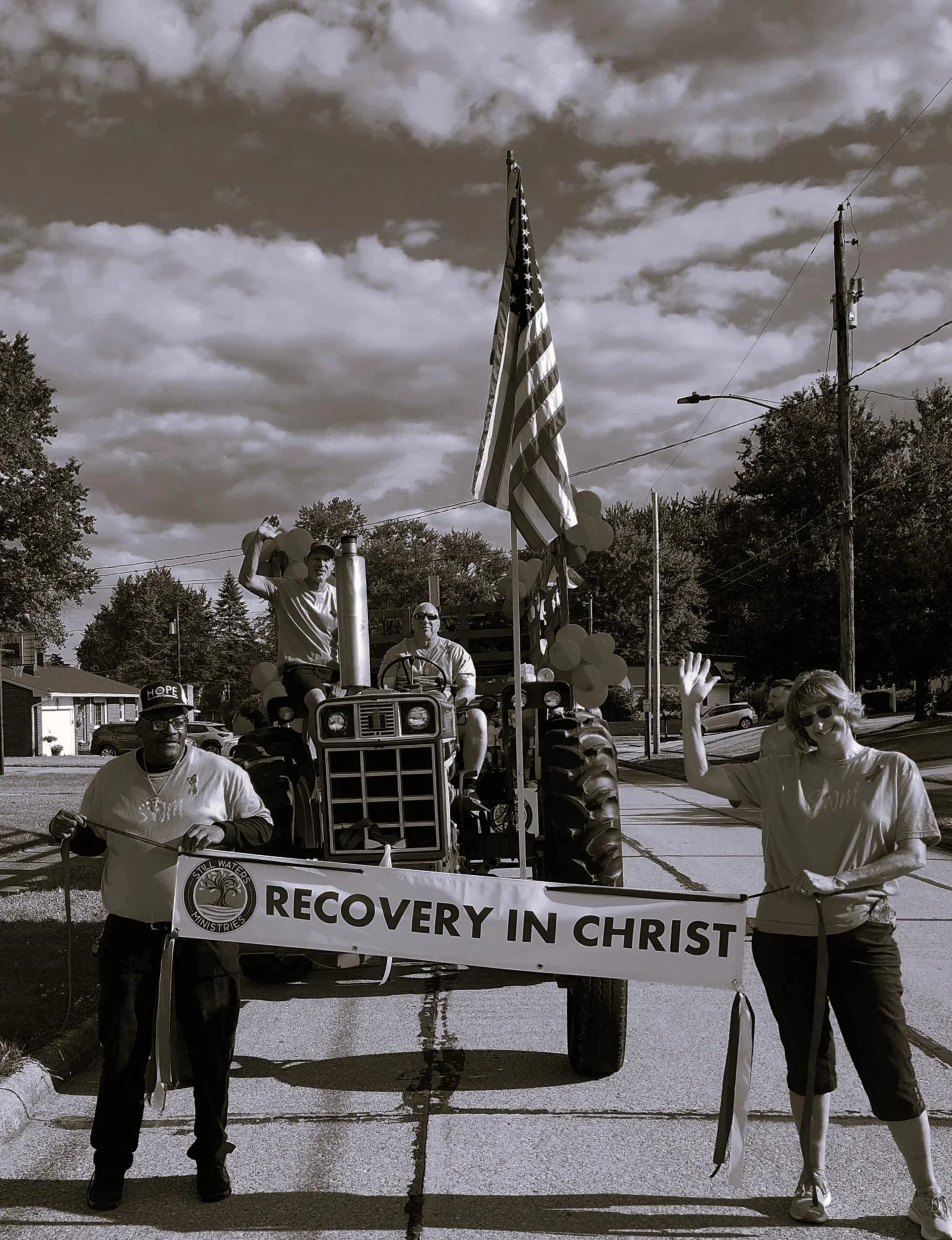 People happily participating in a community parade holding a banner that reads 'Recovery in Christ' with balloons attached to a tractor.