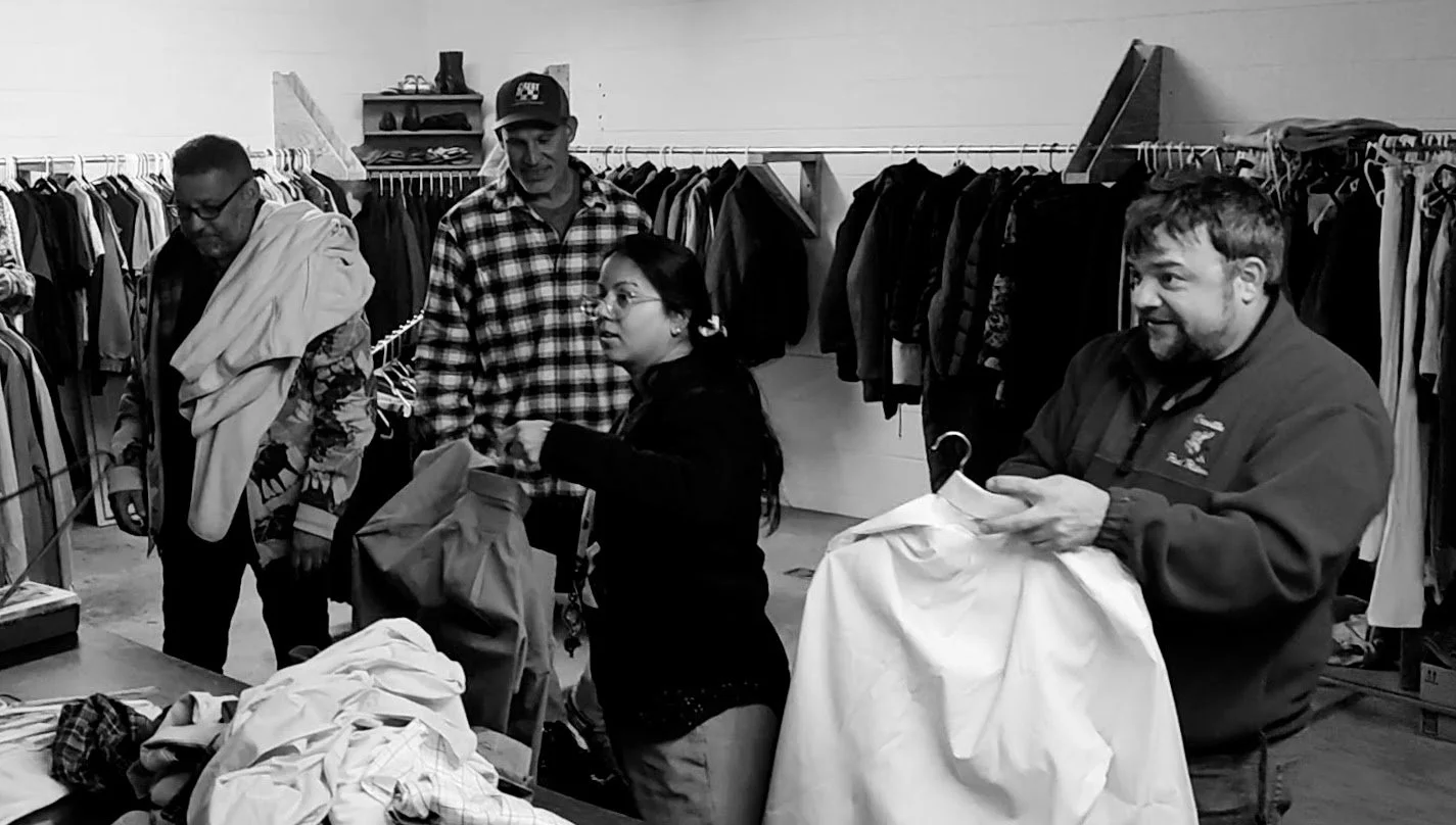 Four people shopping for clothes in a thrift store, with clothing racks in the background.