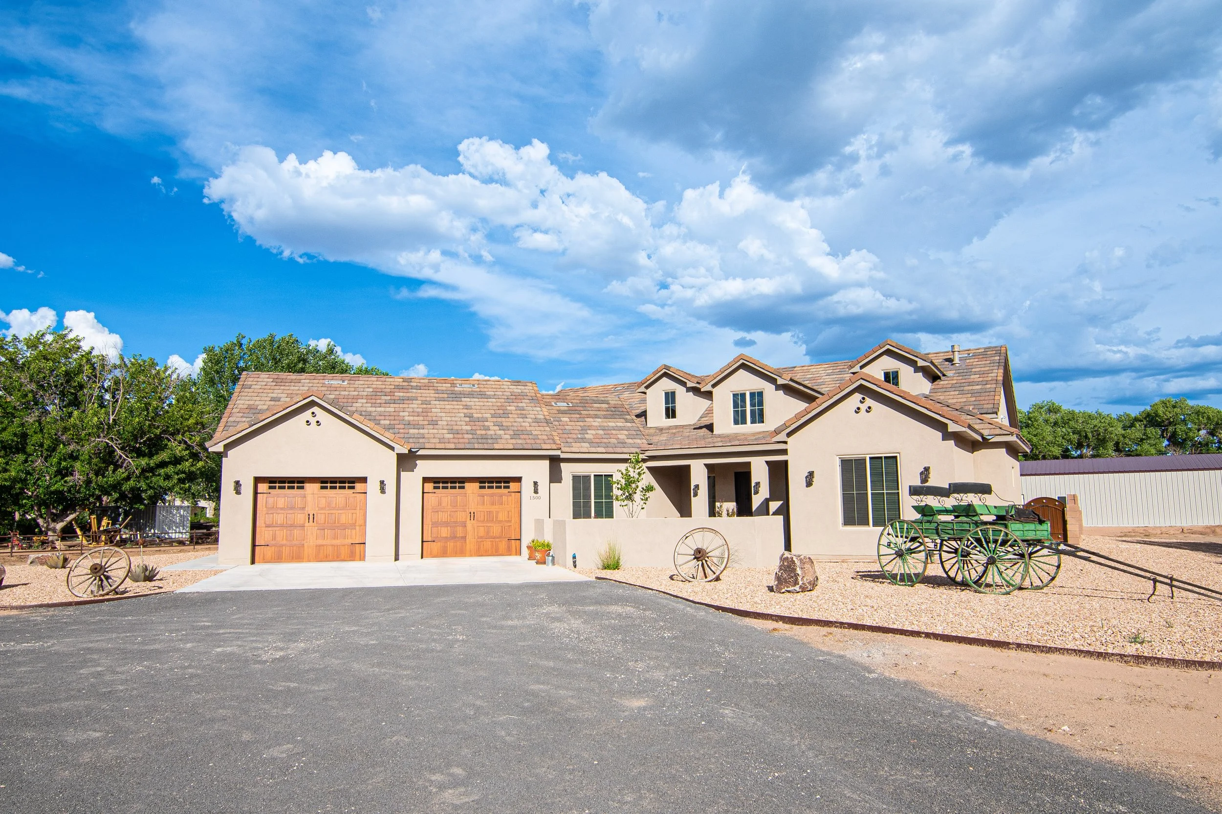 A large, modern house with a two-car garage, white stucco exterior, and a brown tiled roof, situated on a gravel yard with vintage wooden wagons and green landscaping. The sky above is bright blue with white clouds.
