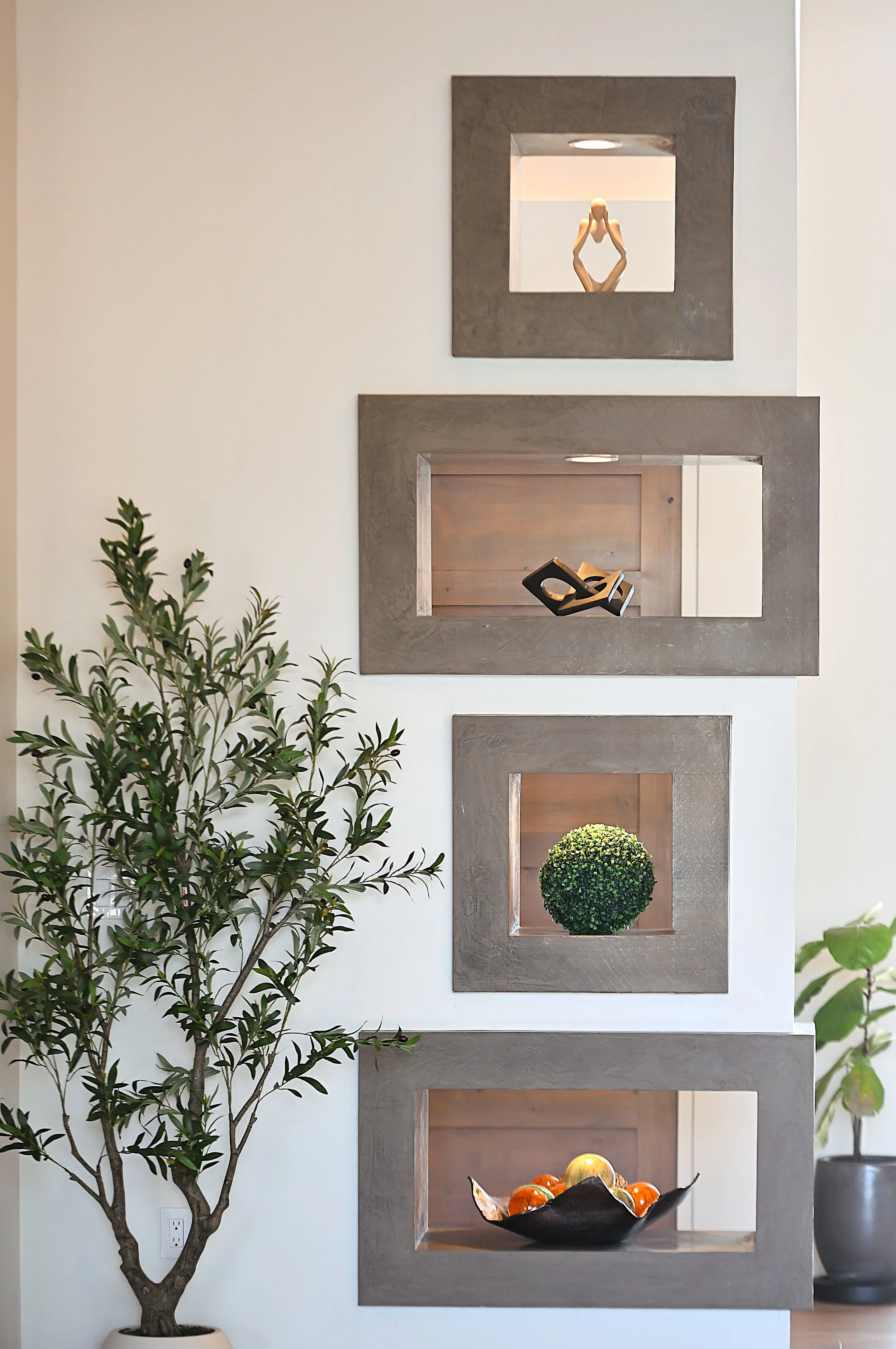 Interior wall with three square framed openings, each containing decorative items: a wooden sculpture of a person, a black abstract sculpture, and a potted round green plant. To the left, a tall indoor tree with green leaves, and to the right, part of a gray pot is visible.