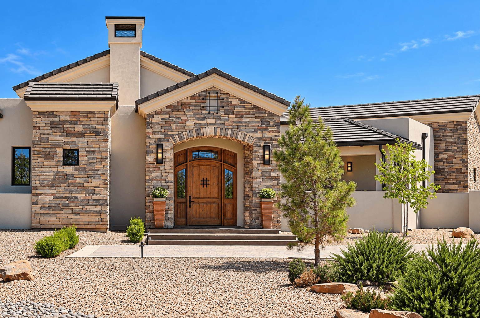 Front view of a modern house with a combination of brick and stucco exterior, a wooden front door, and landscaped yard with small trees and rocks.