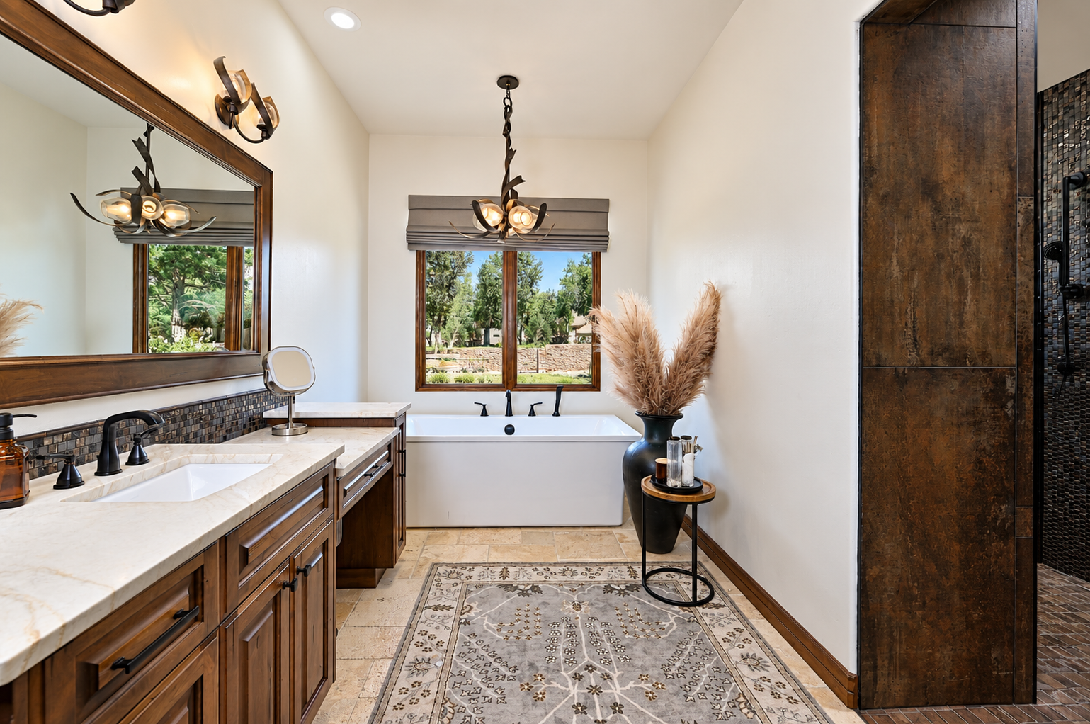 Bathroom with double vanity, large mirror, bathtub near window, chandelier, beige carpet, black vase with pampas grass, and brown cabinets.