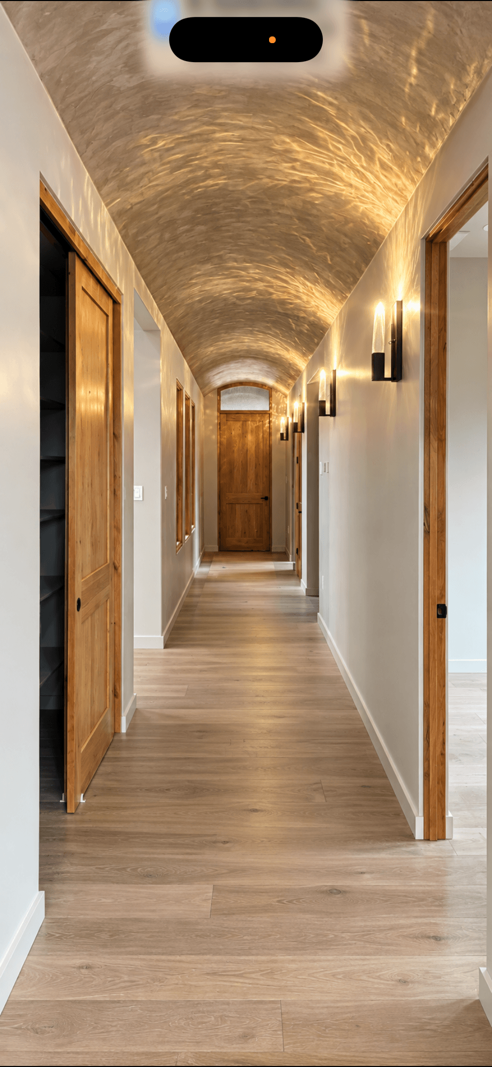 A hallway with wooden trim, light wooden flooring, a textured arched ceiling, and wall-mounted lights.