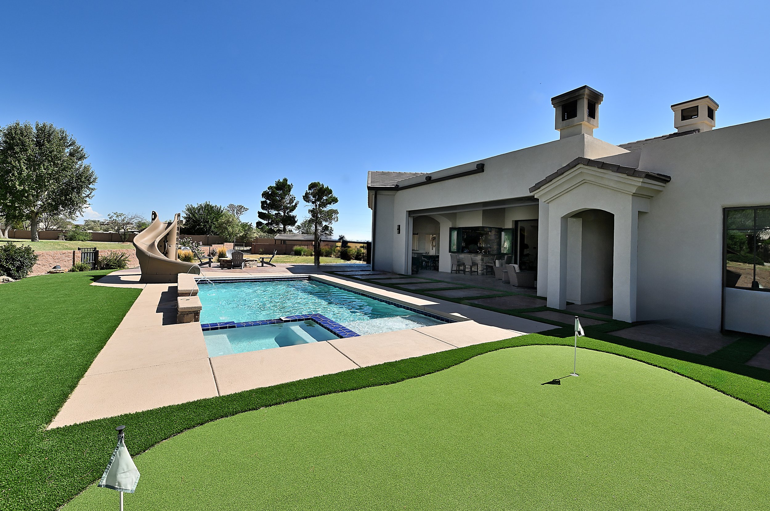 Backyard with a swimming pool, a lawn, and a house with patio furniture under a clear blue sky.