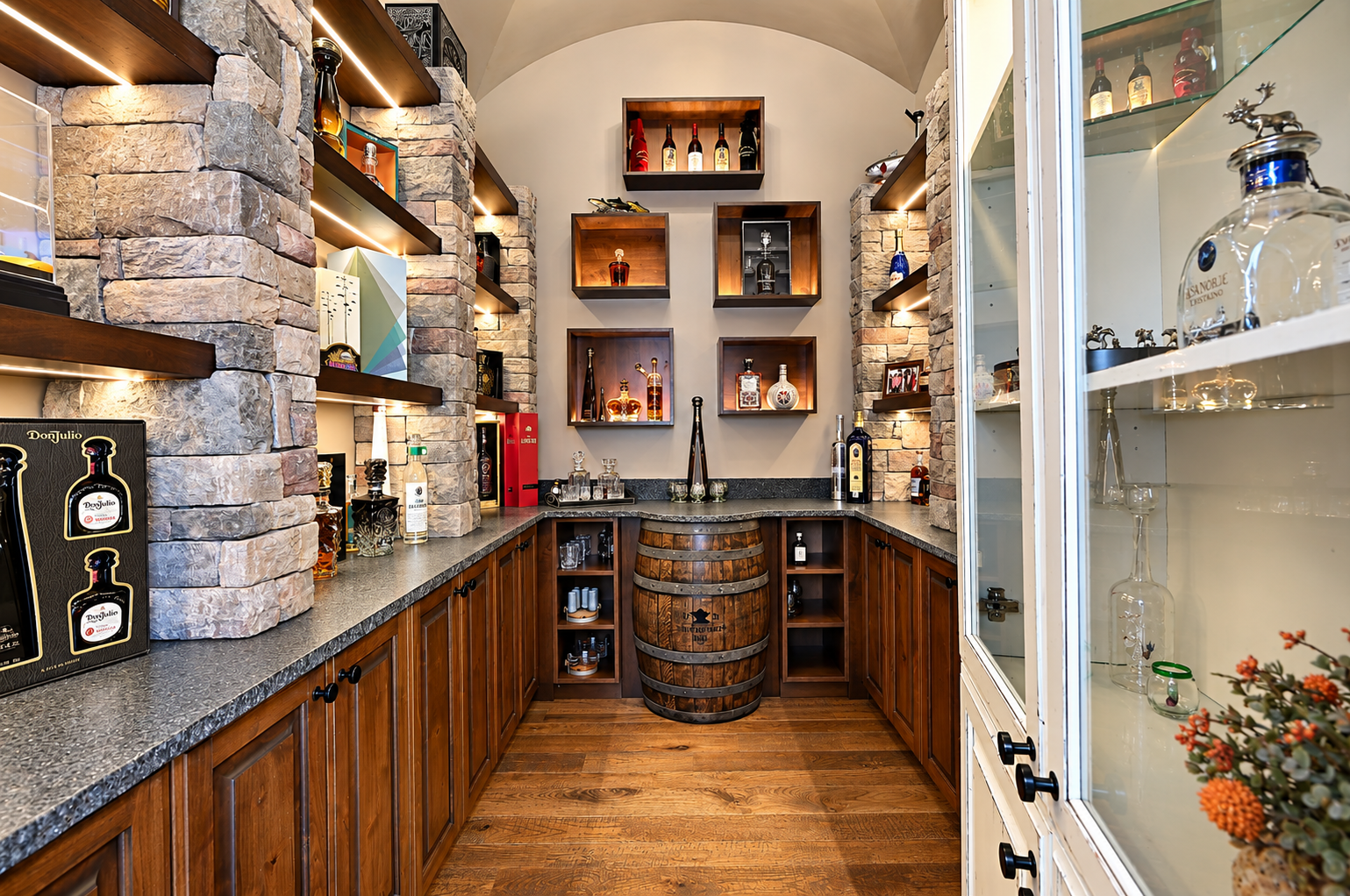 A home bar with stone walls, wooden shelves, and glassware display, featuring bottles of alcohol and decorative items, with a wine barrel in the center.
