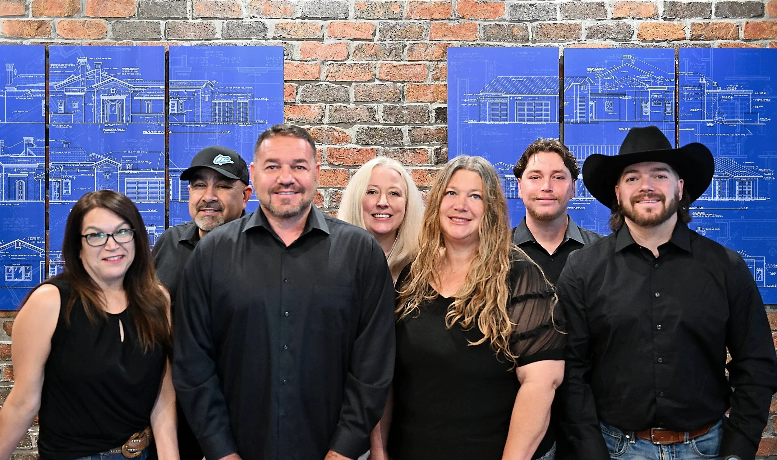 The team at Quality Builders New Mexico stand together in front of architectural blueprints displayed on a brick wall.