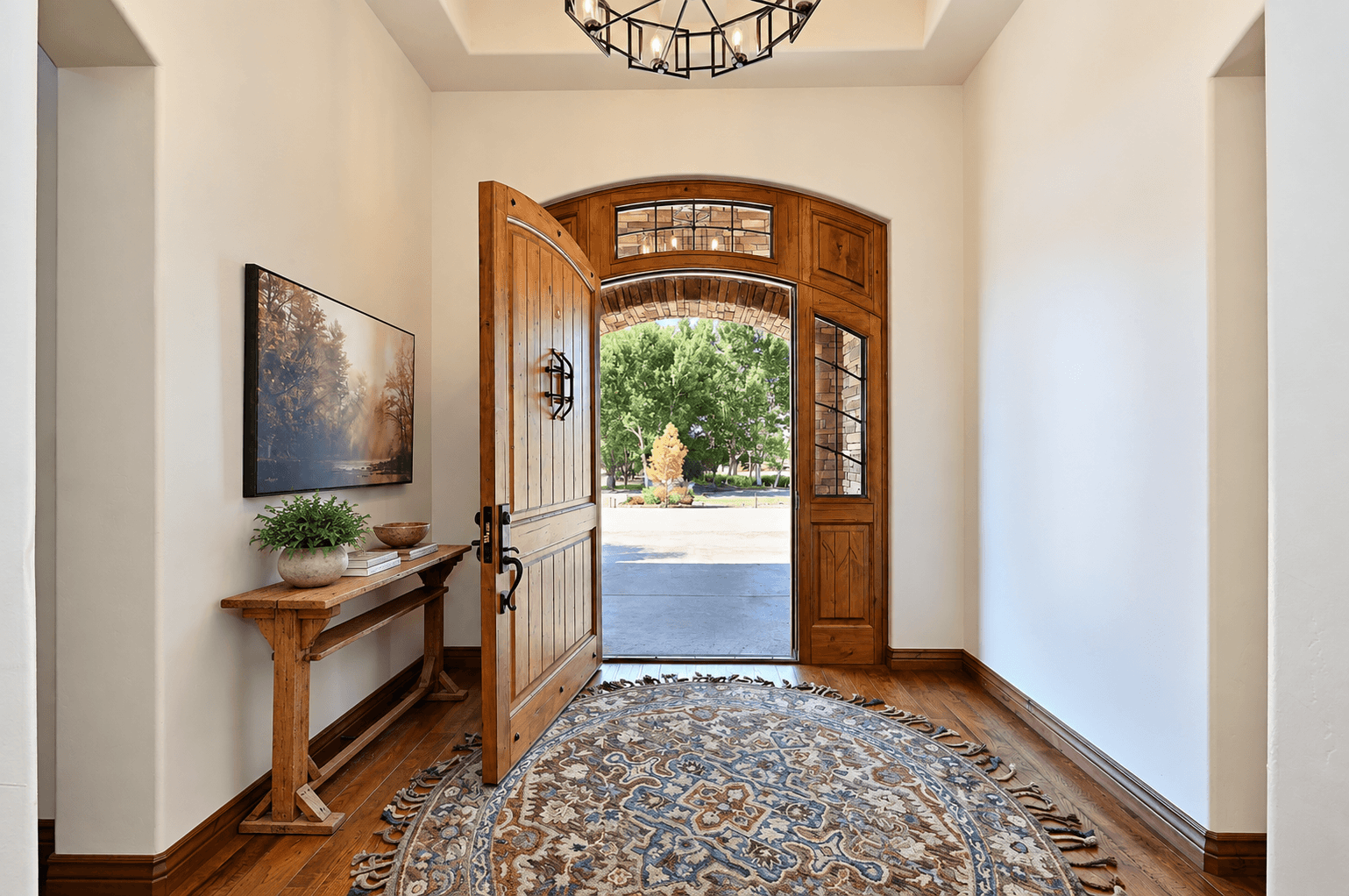 View of a wooden front door opening to a sunny street with trees and a small shrub, inside an entryway with hardwood floors, a decorative rug, and a side table with potted plant and decor.