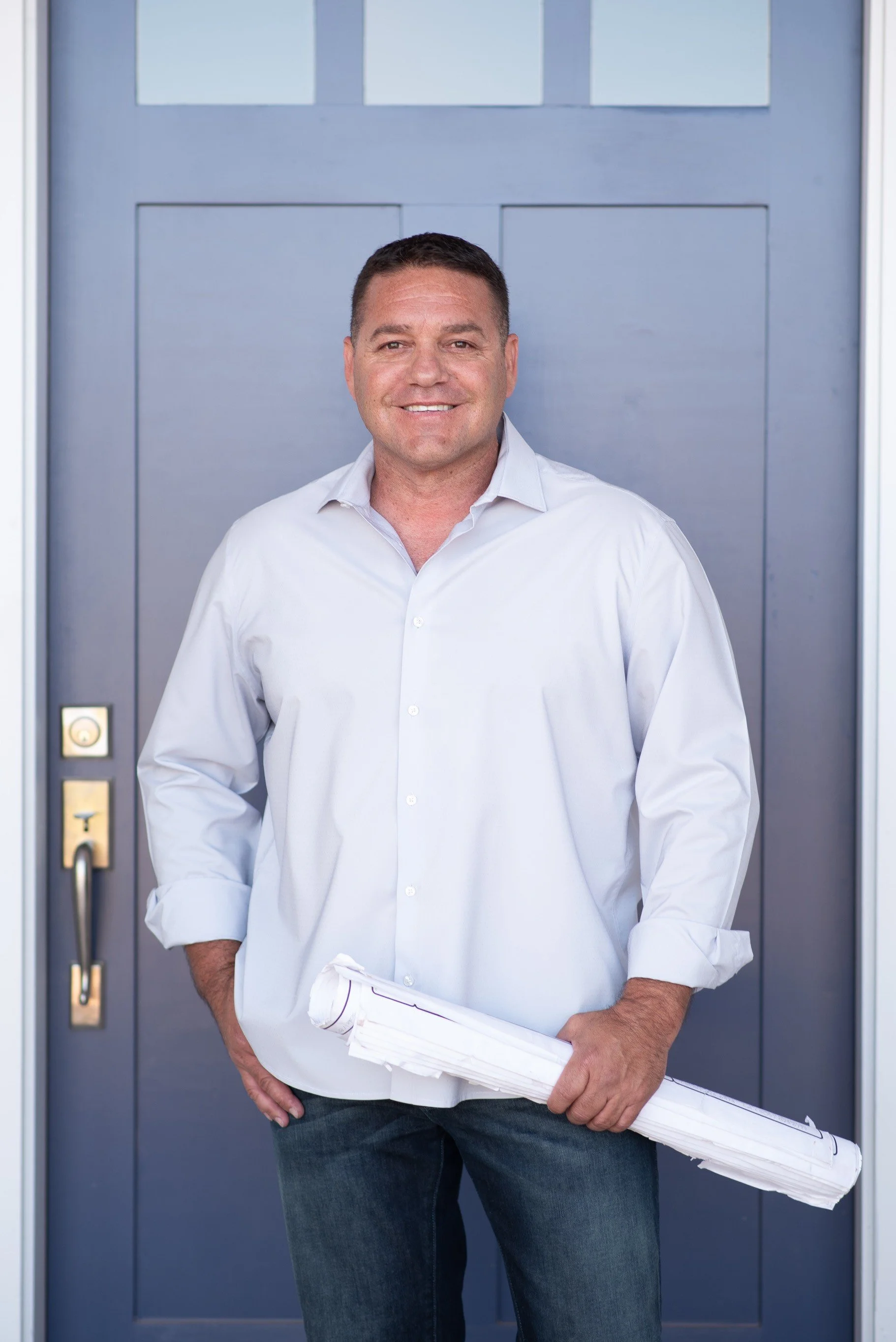Mike Martinez, Owner of Quality Builders, stands in front of a door holding blueprints.