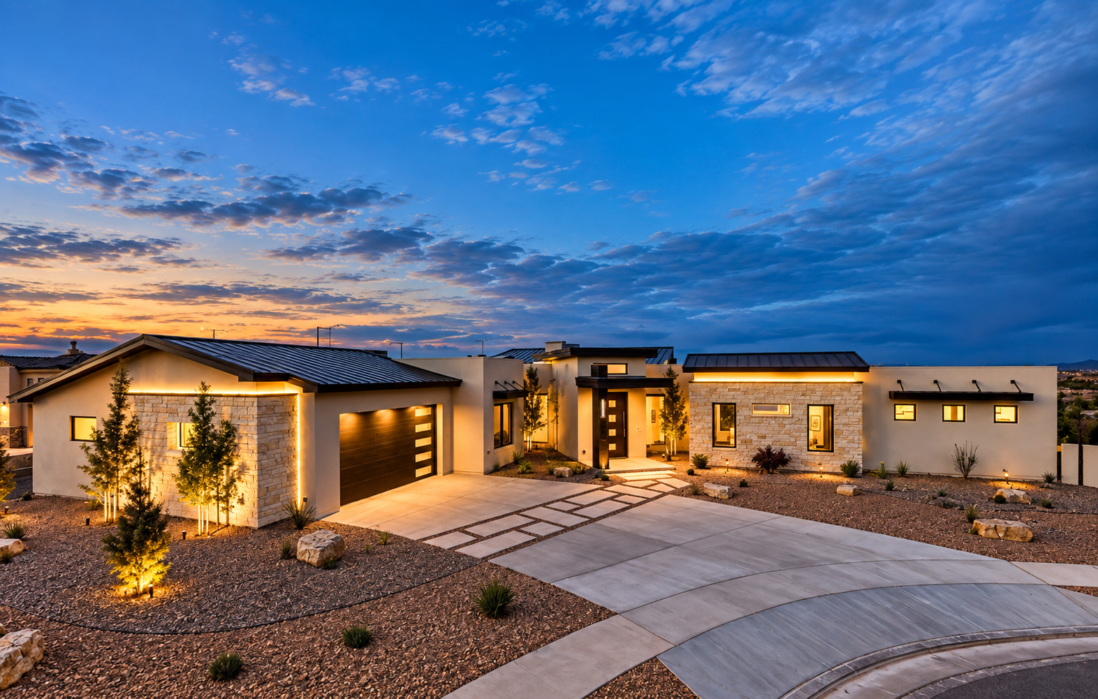 Modern house at dusk with illuminated exterior lighting, a wide driveway, and a minimalist landscape with small trees and rocks.