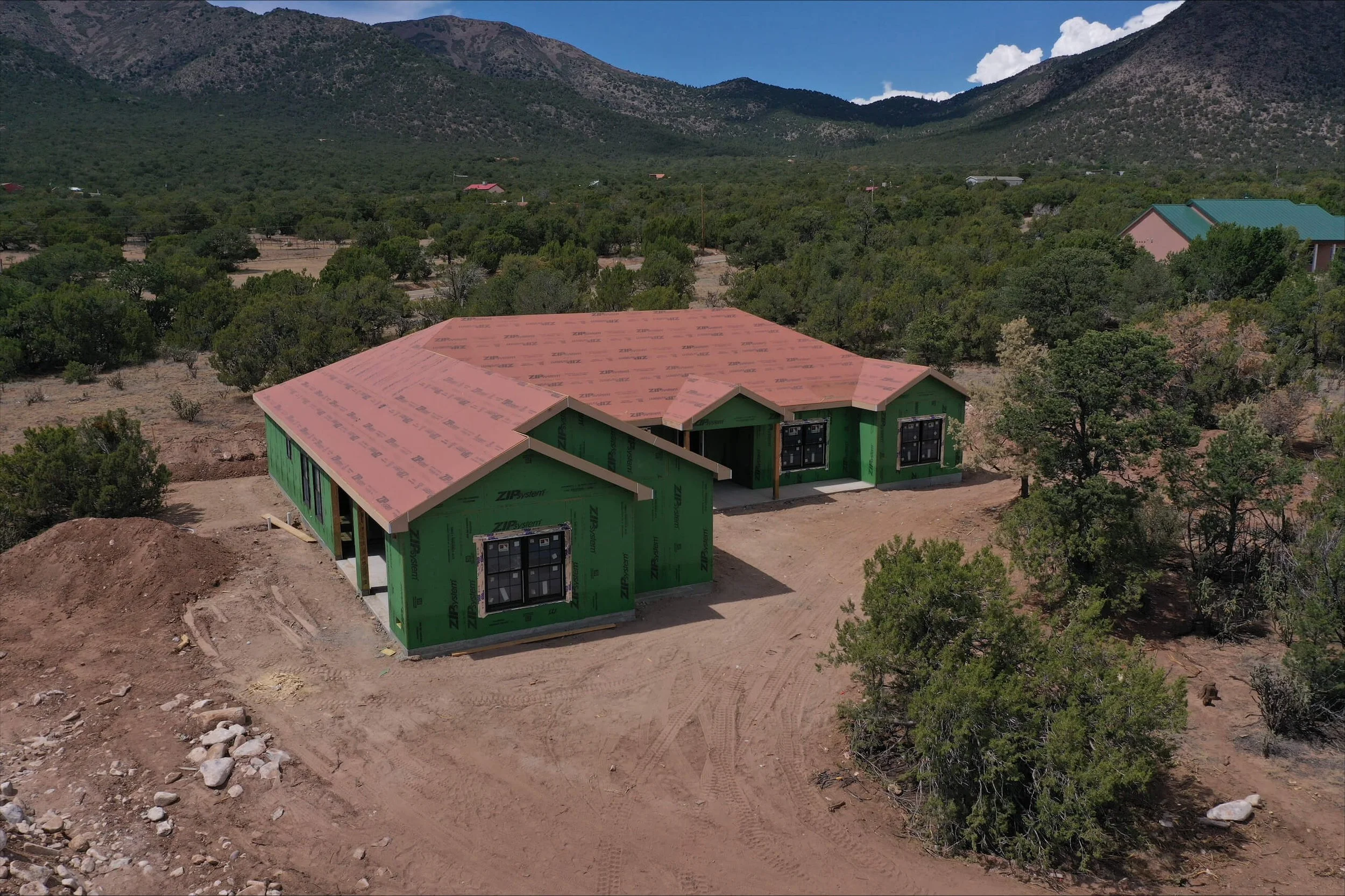 A house under construction with green exterior walls and a pinkish-brown roof, surrounded by dirt and rocks, with a backdrop of green trees, mountains, and a partly cloudy sky.