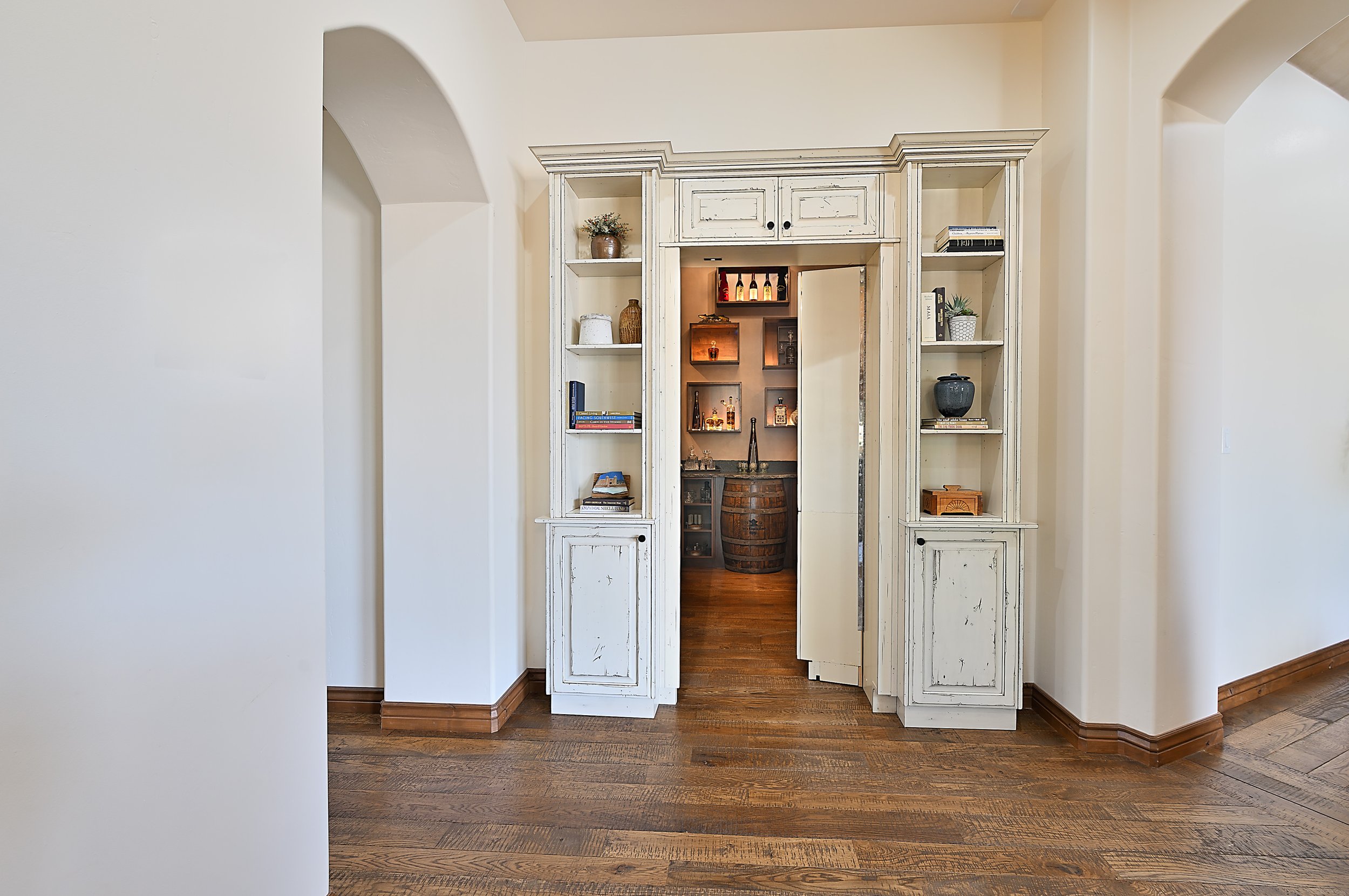 White built-in bookshelf with decorative items and books, opening into a room with a barrel, on a wooden floor with light-colored walls and arches.