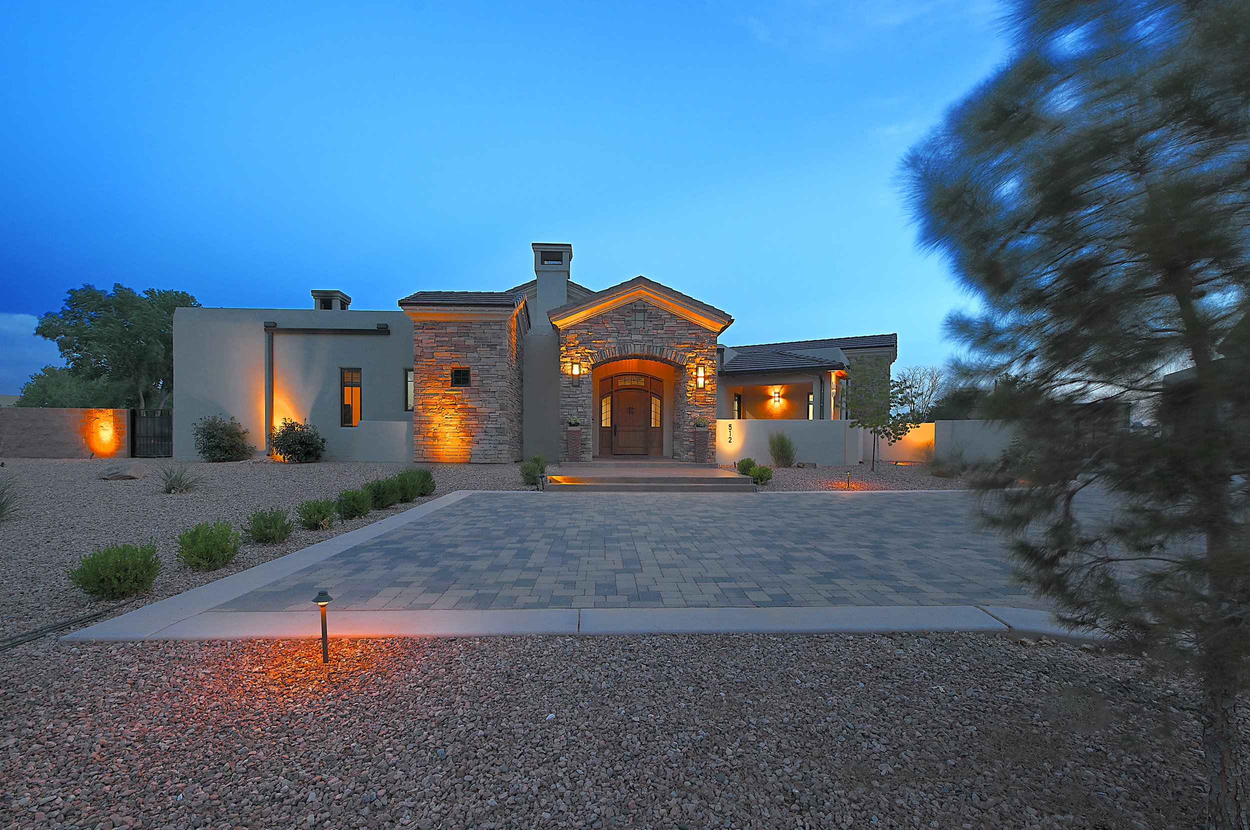 Modern house with stone and stucco exterior, lit entrance, surrounded by desert landscape with small bushes, trees, and gravel yard, illuminated by exterior lights at dusk.