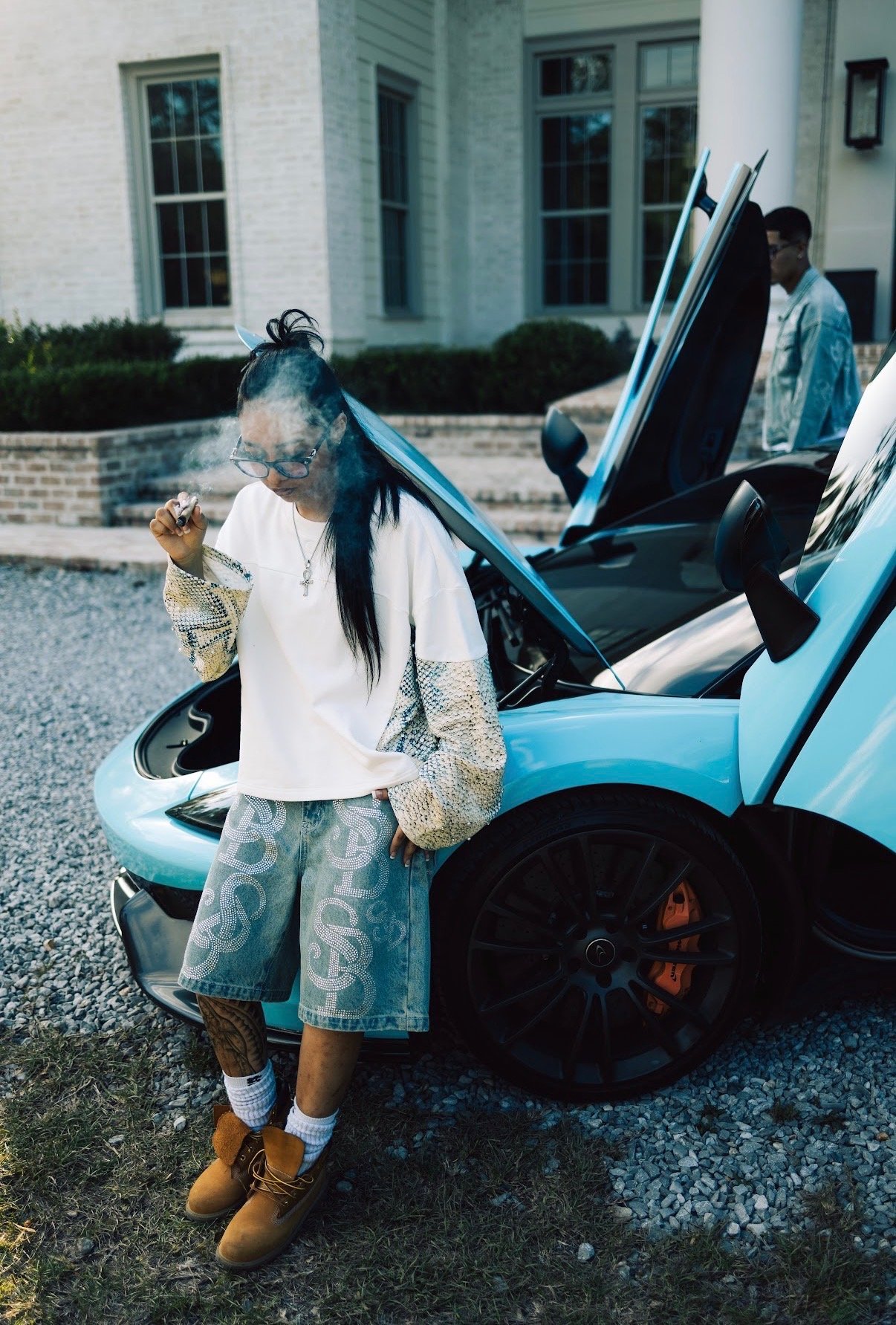 A woman is standing next to a light blue sports car with its doors open, smoking a cigar and dressed in casual clothing, with two men and a large white house in the background.