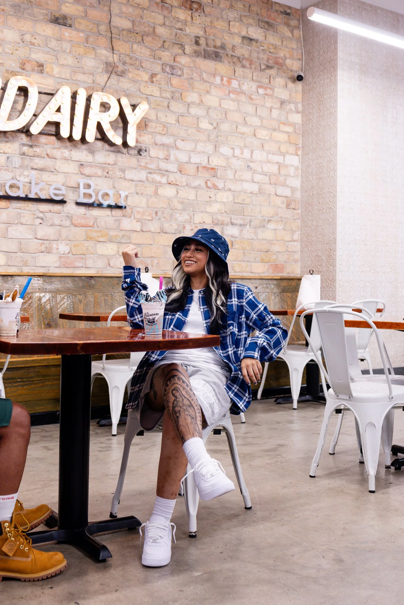 A young woman with long blonde and black hair, wearing a blue bucket hat, a blue plaid shirt, white shorts, and white sneakers, sitting in a casual cafe or ice cream shop with a brick wall behind her. She is smiling and flexing her arm muscles, with a tattoo on her left leg visible. There are cups of ice cream or milkshakes on the table.