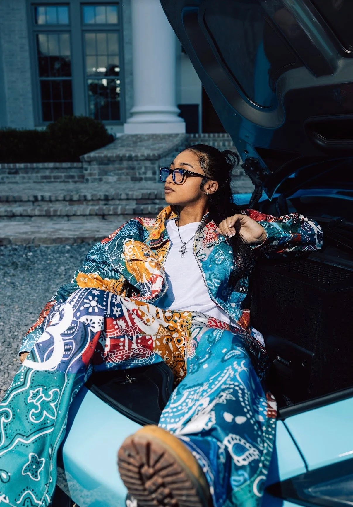 A young woman with glasses and long dark hair, dressed in colorful, patterned clothing, relaxing against the open trunk of a car parked outside a house with brick steps and large windows.