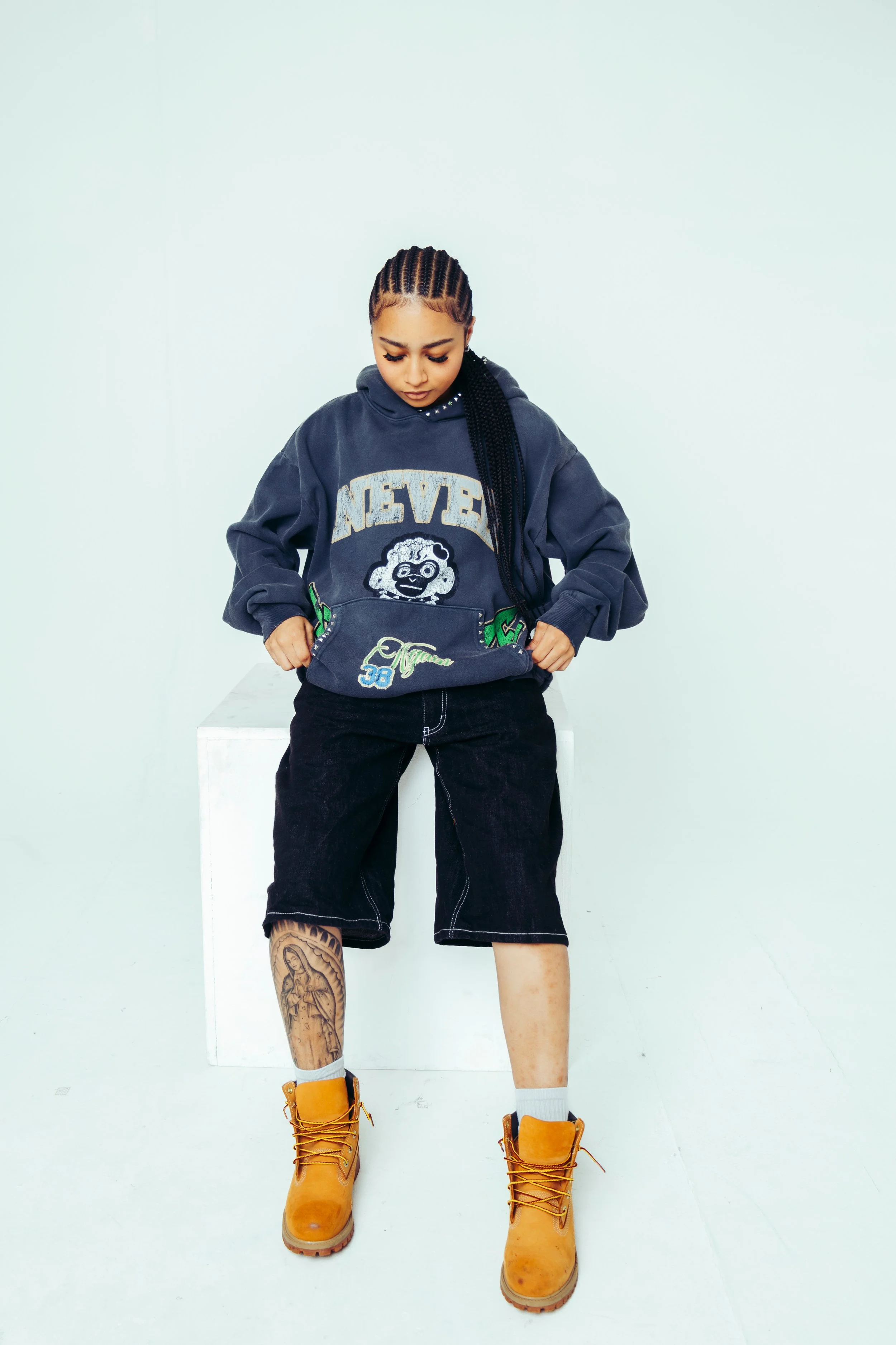 A woman standing in front of a plain light-colored wall, wearing a dark sweatshirt with graphics and text, black denim shorts, yellow boots, and showing a tattoo on her left shin. She has braided hair and looks down.