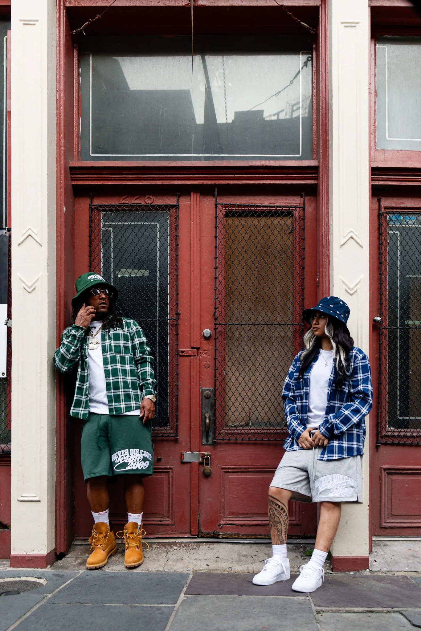 Two young adults standing in front of a red door with black security grates, wearing casual streetwear and bucket hats. The man on the left wears a green plaid shirt, green shorts, and brown boots, while the woman on the right wears a blue plaid shirt, gray shorts, and white sneakers.
