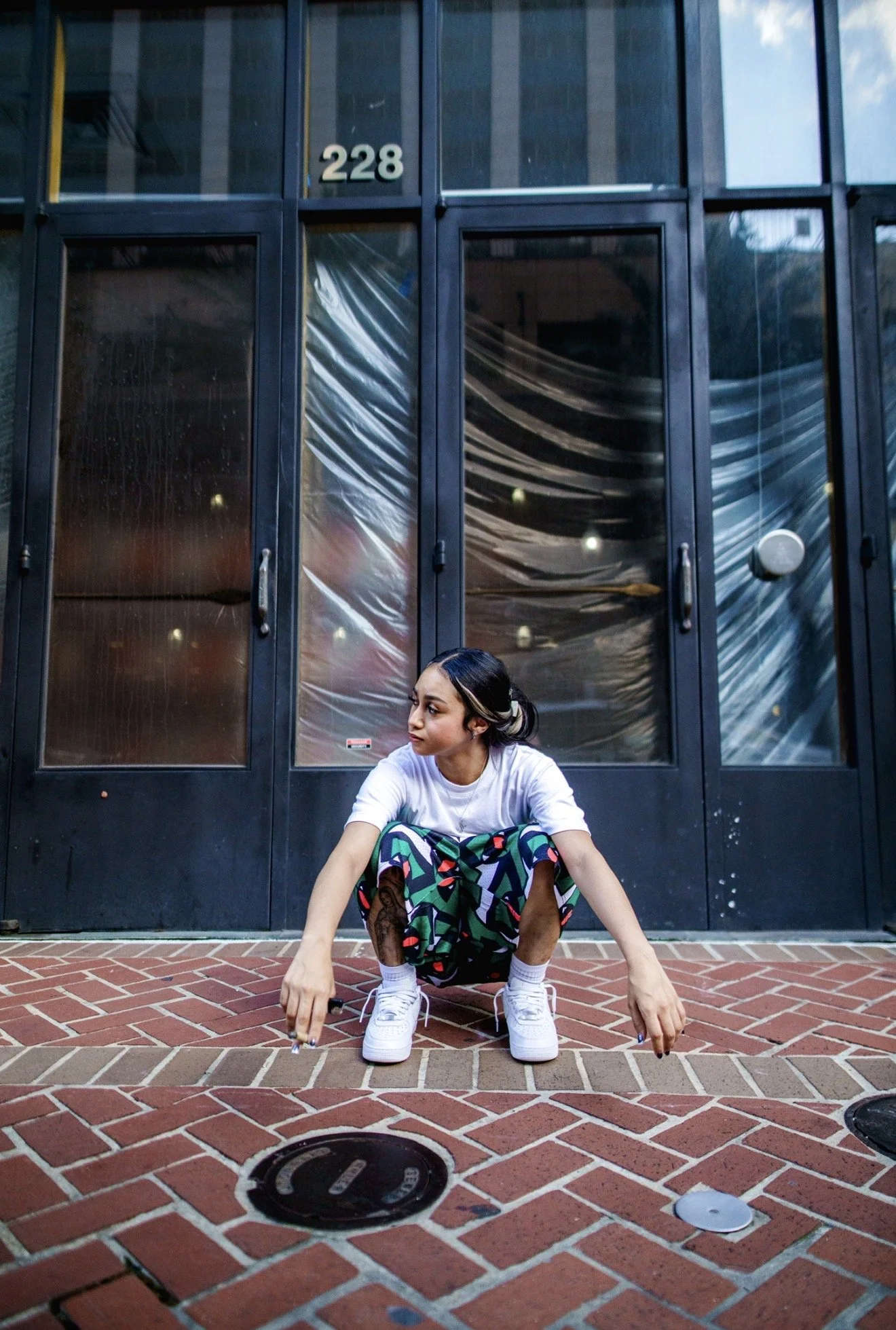A young woman with dark hair, sitting in a crouched position on a brick sidewalk in front of a black door, wearing colorful patterned pants, white sneakers, a white T-shirt, and holding a skateboard.