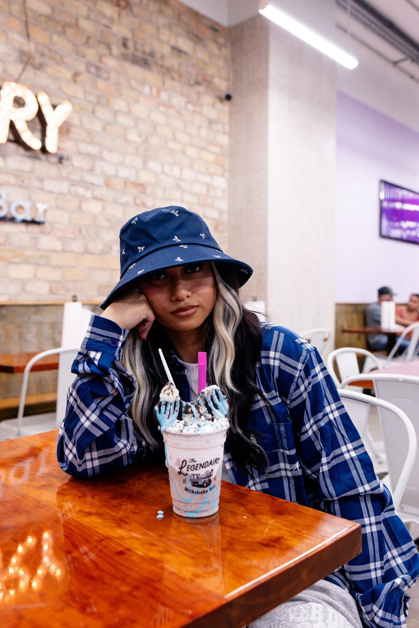 A young woman with long hair wearing a blue bucket hat and a blue plaid shirt sitting at a wooden table in an ice cream shop. She has her head resting on her hand and looks at the camera. In front of her is a large milkshake topped with blue and white icing, whipped cream, and two blue wafer sticks. The shop interior includes a brick wall, a neon sign, and people seated at tables in the background.
