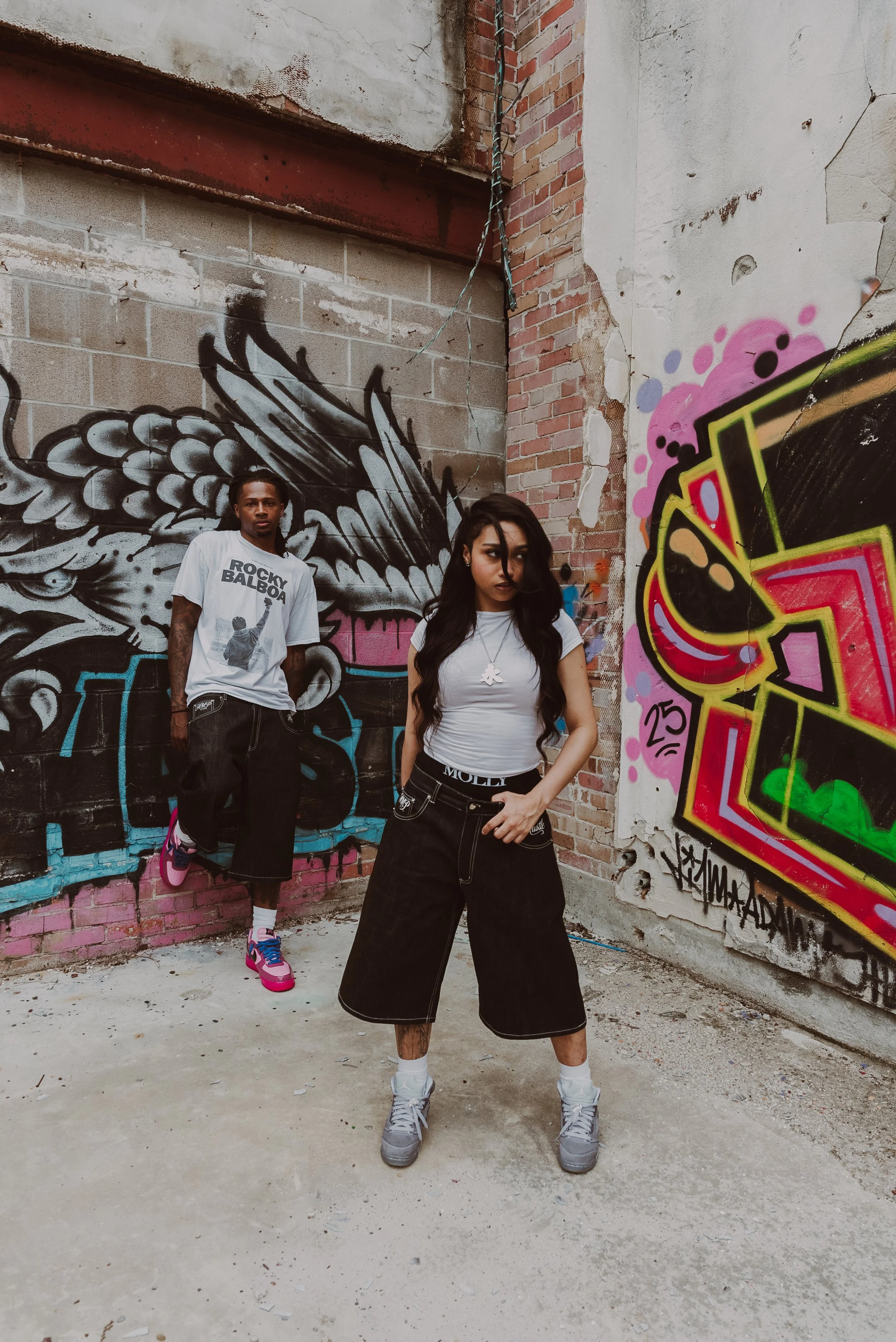 Two young adults, a man and a woman, posing in front of graffiti art on an urban brick wall. The man stands with one foot on the ground and the other resting on the wall, wearing a white graphic T-shirt, black shorts, and colorful sneakers. The woman stands with her hands in her pockets, wearing a white t-shirt, black shorts, and gray sneakers, with long dark hair partially covering her face.