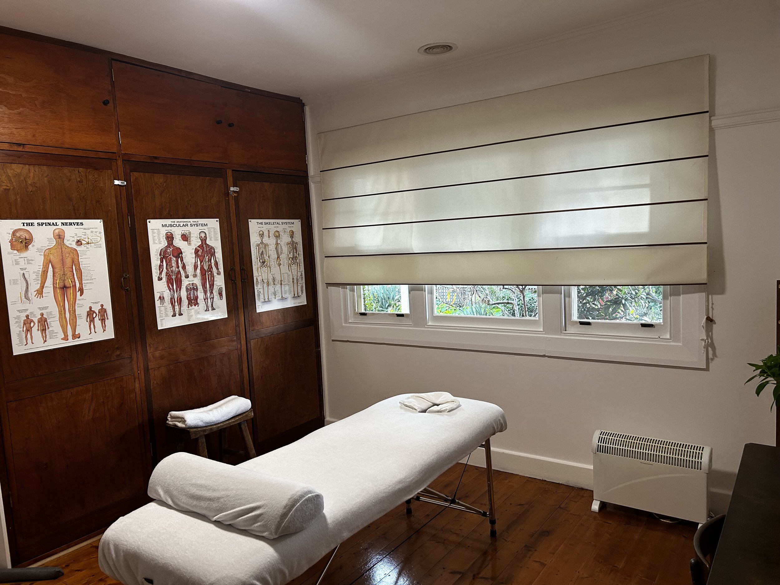 A massage therapy room with a massage table covered in a white sheet, rolled towels on a small stool, wooden cabinets with anatomical posters, and a large window with shutters letting in natural light.