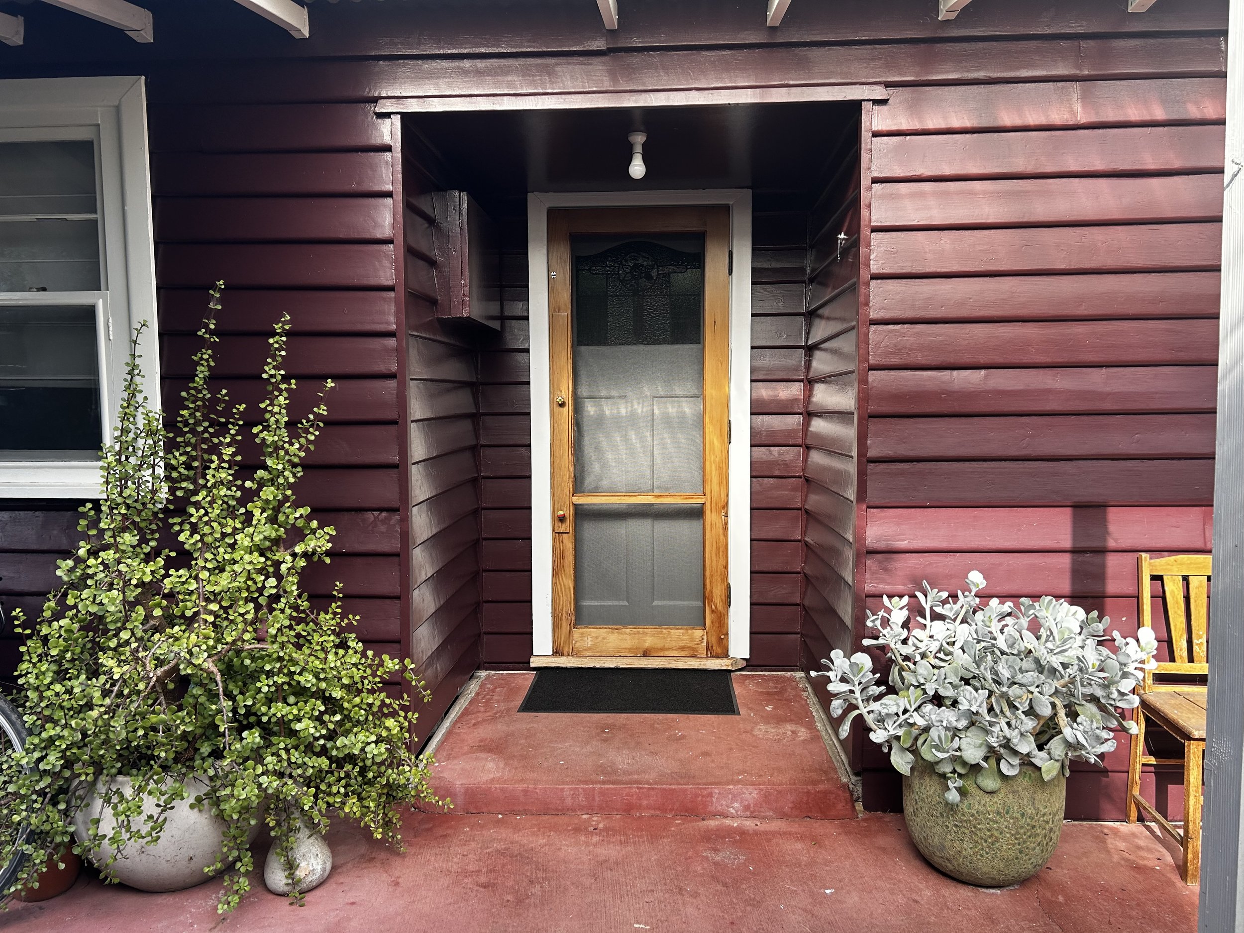 The front entrance of a house with a wooden door and a screen door, surrounded by plants in pots and a wooden bench on the porch.