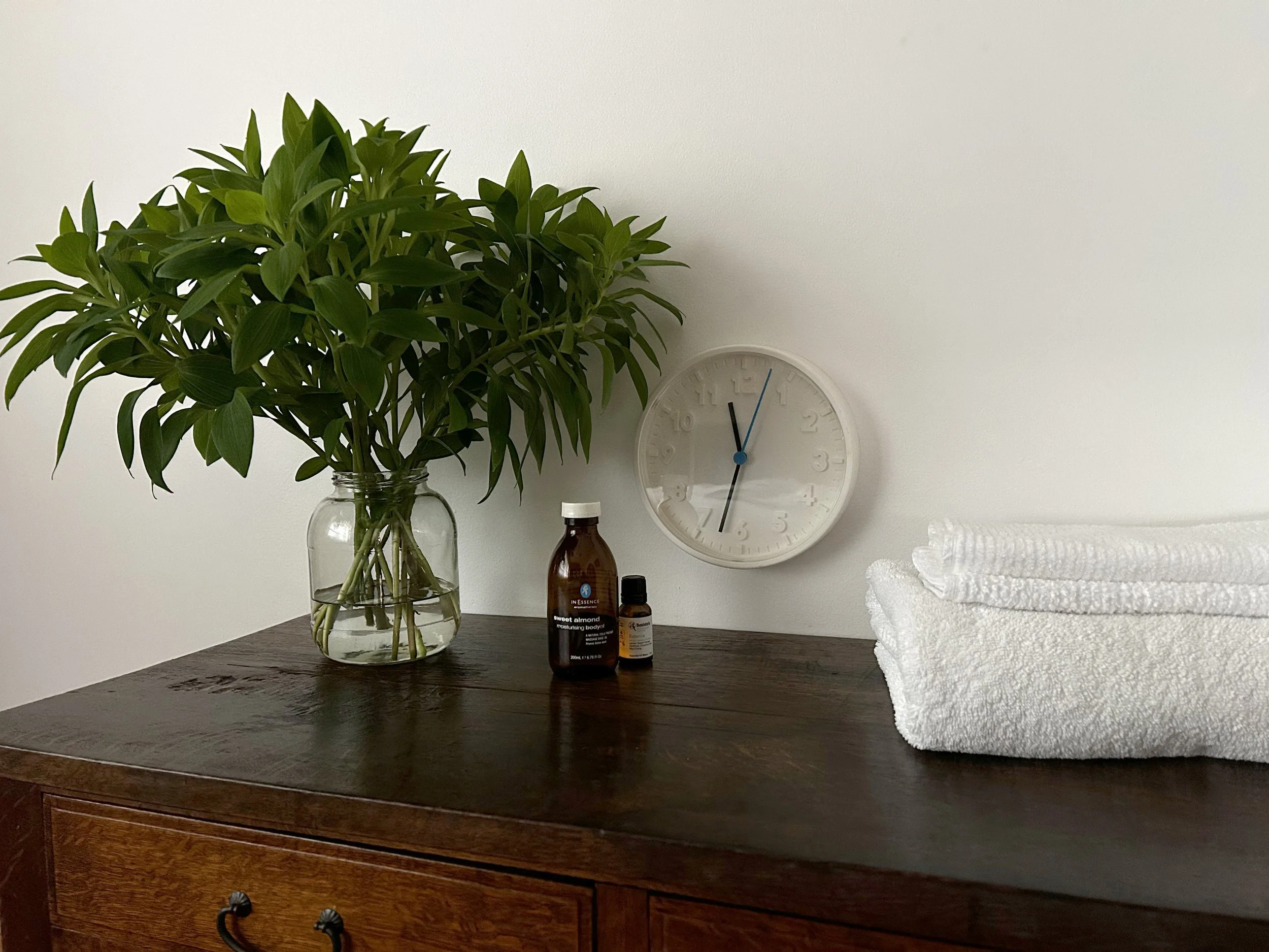 A wooden dresser with a glass vase holding green leafy plant, two small bottles of emulsion and vitamins, folded white towels, and a white wall clock on a white wall.
