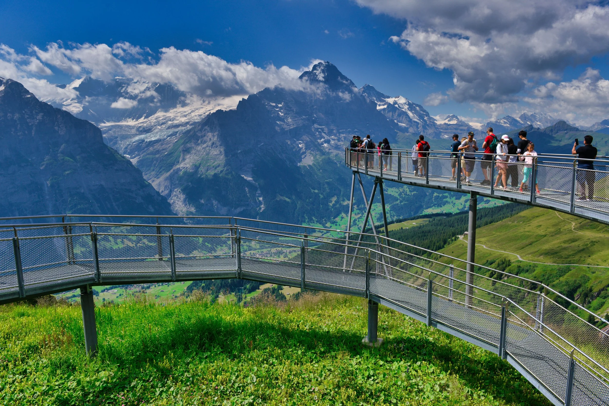 A group of tourists walking on a suspended skywalk with mountain scenery, snow-capped peaks, and a partly cloudy sky in the background.