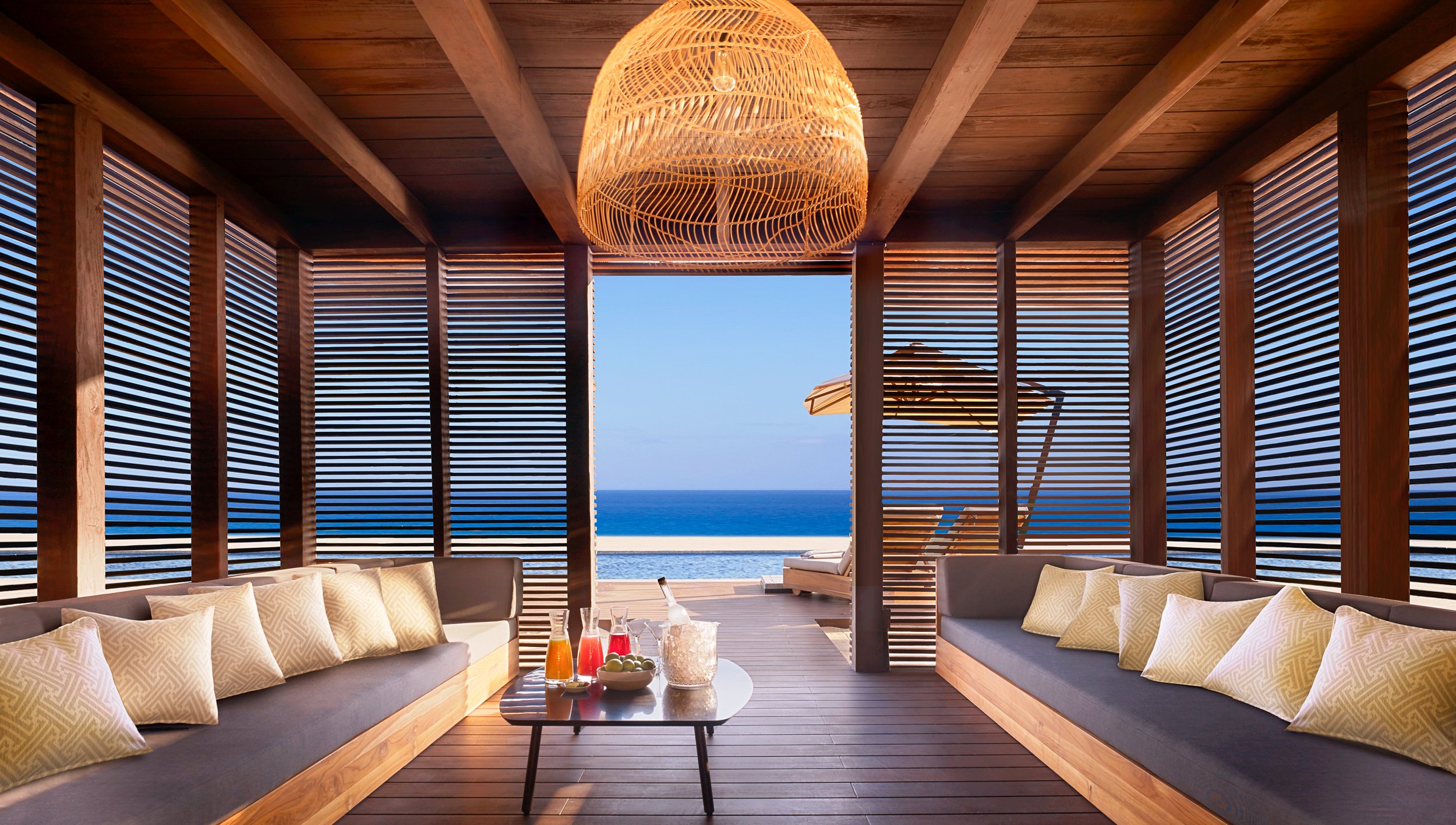 Beachside outdoor lounge area with wooden slats, cushioned seating, a woven pendant lamp, and a view of the ocean and sky in the background.