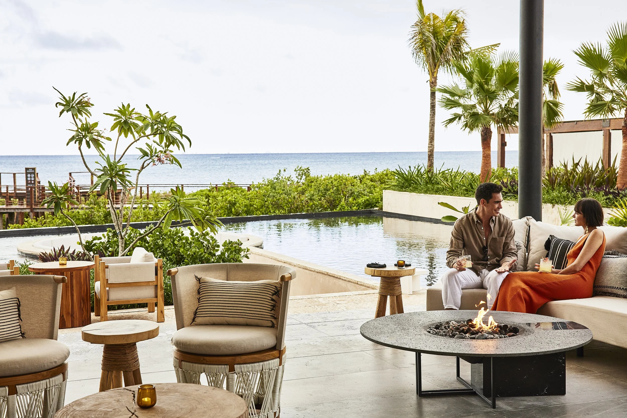 Relaxed outdoor seating area overlooking the ocean, with two people enjoying drinks around a fire pit, surrounded by palm trees and lush greenery.